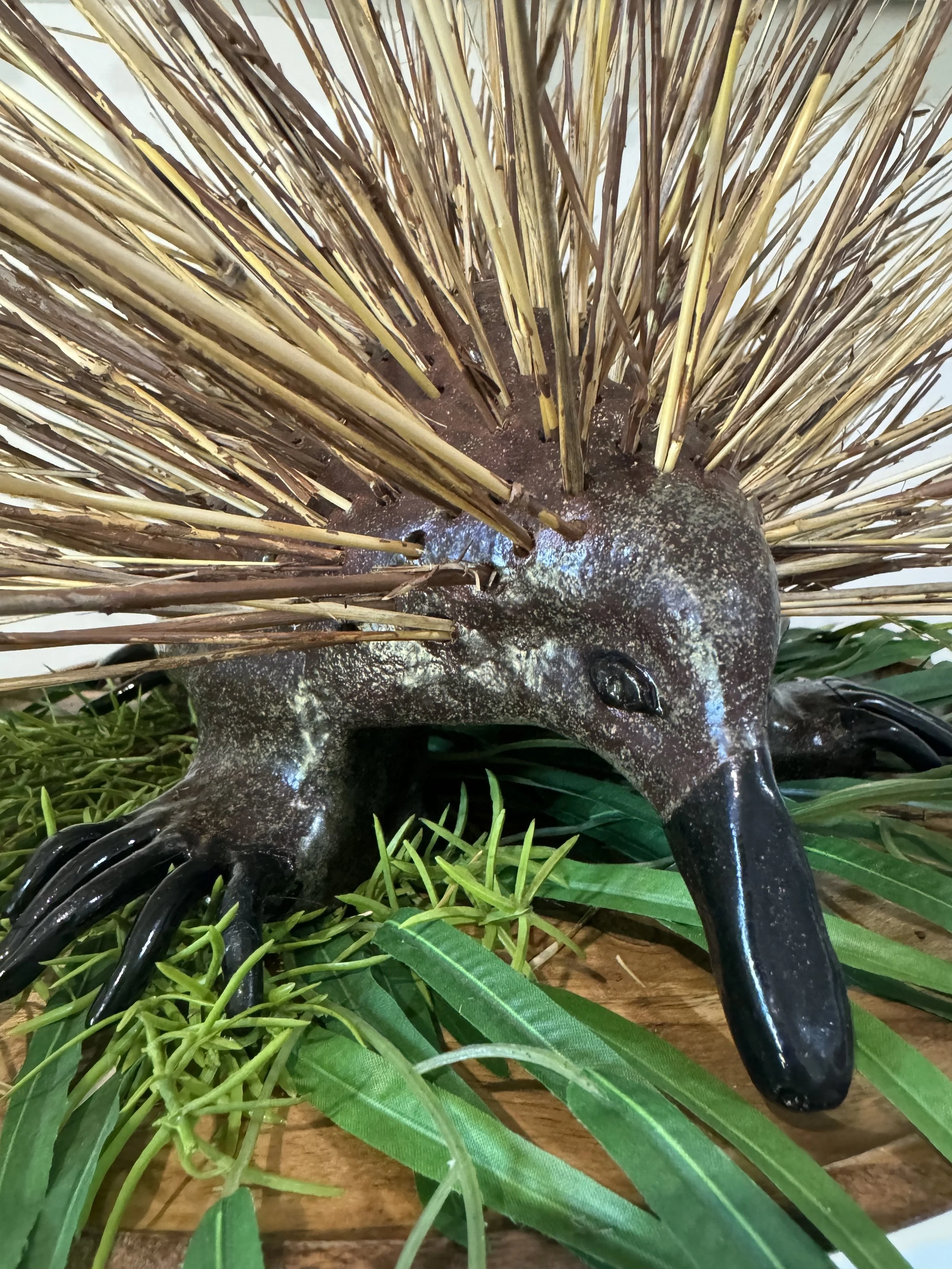 A decorative sculpture of a hedgehog-like creature with a dark body, a large nose, and spikes made of straw or similar material, surrounded by green leaves and grass.