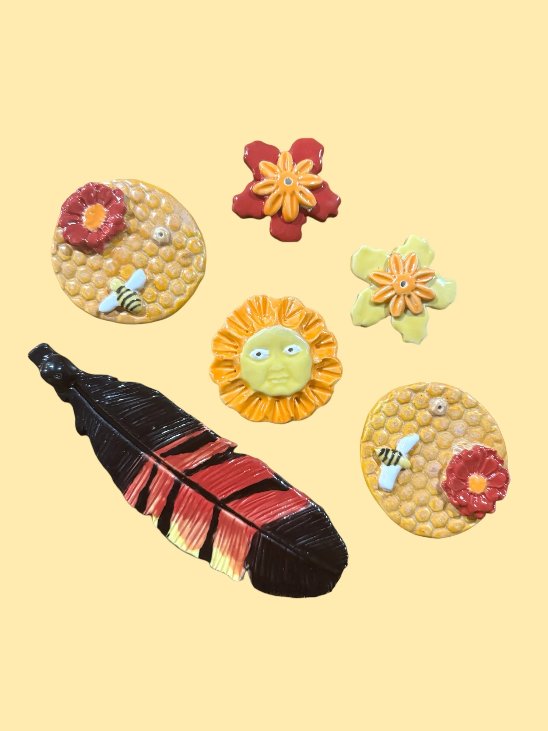 Colorful ceramic art pieces including flowers, a face, honeycomb pattern, a bee, a large feather, and a sun with a face, displayed against a light yellow background.