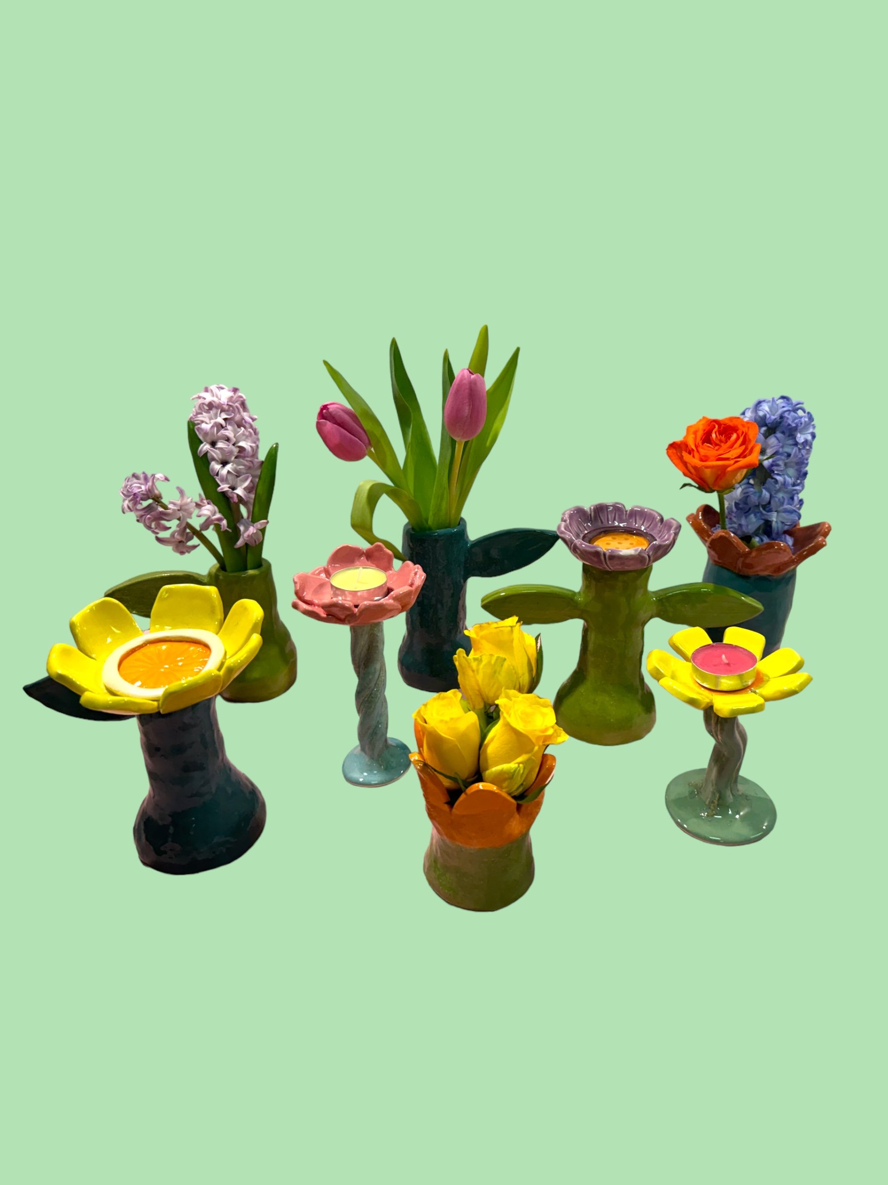 Colorful ceramic flower vases with artificial flowers and candles on a light green background.