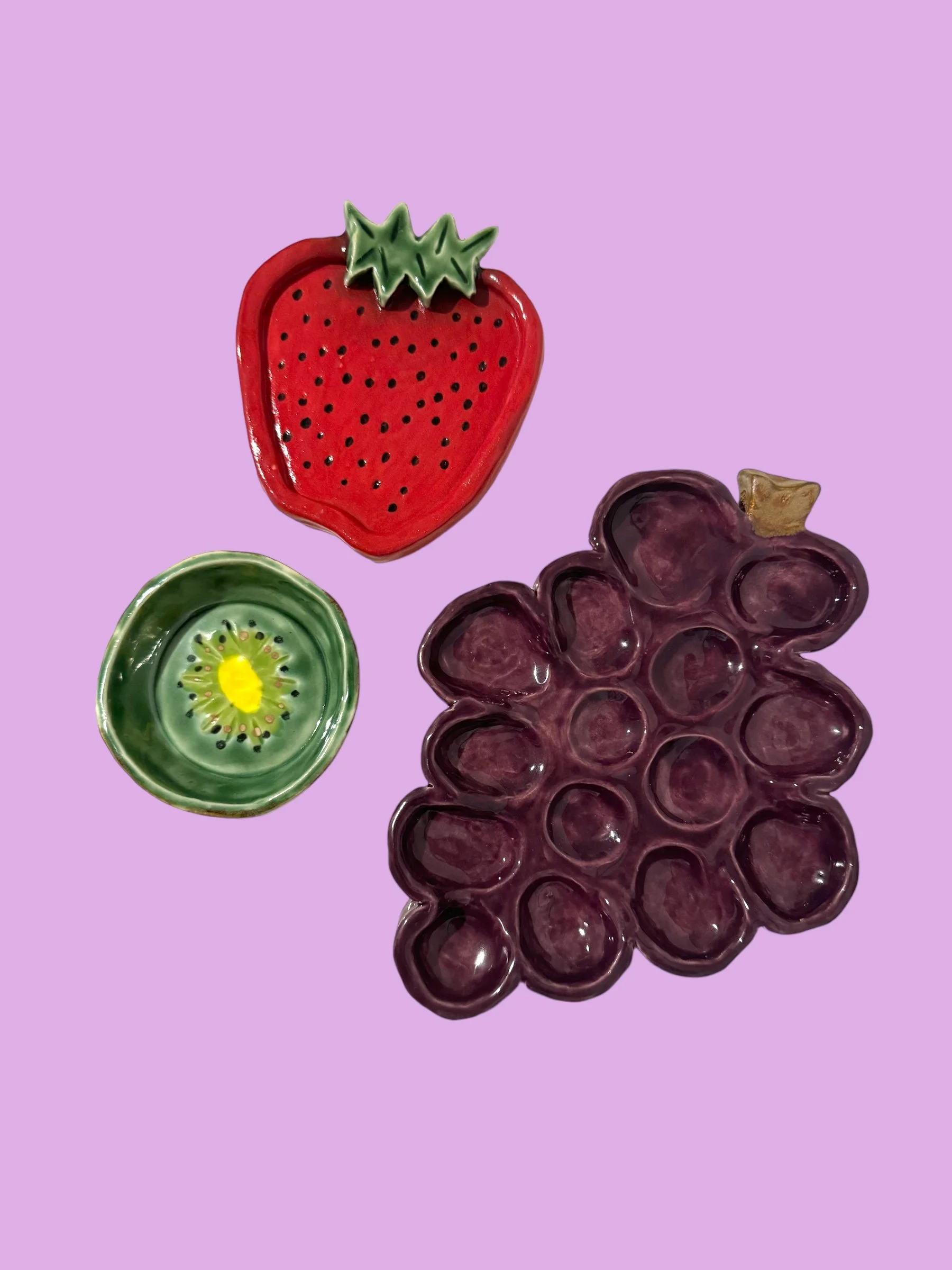 Decorative ceramic dishes shaped like a strawberry, kiwi, grapes, and a small bowl on a purple background.