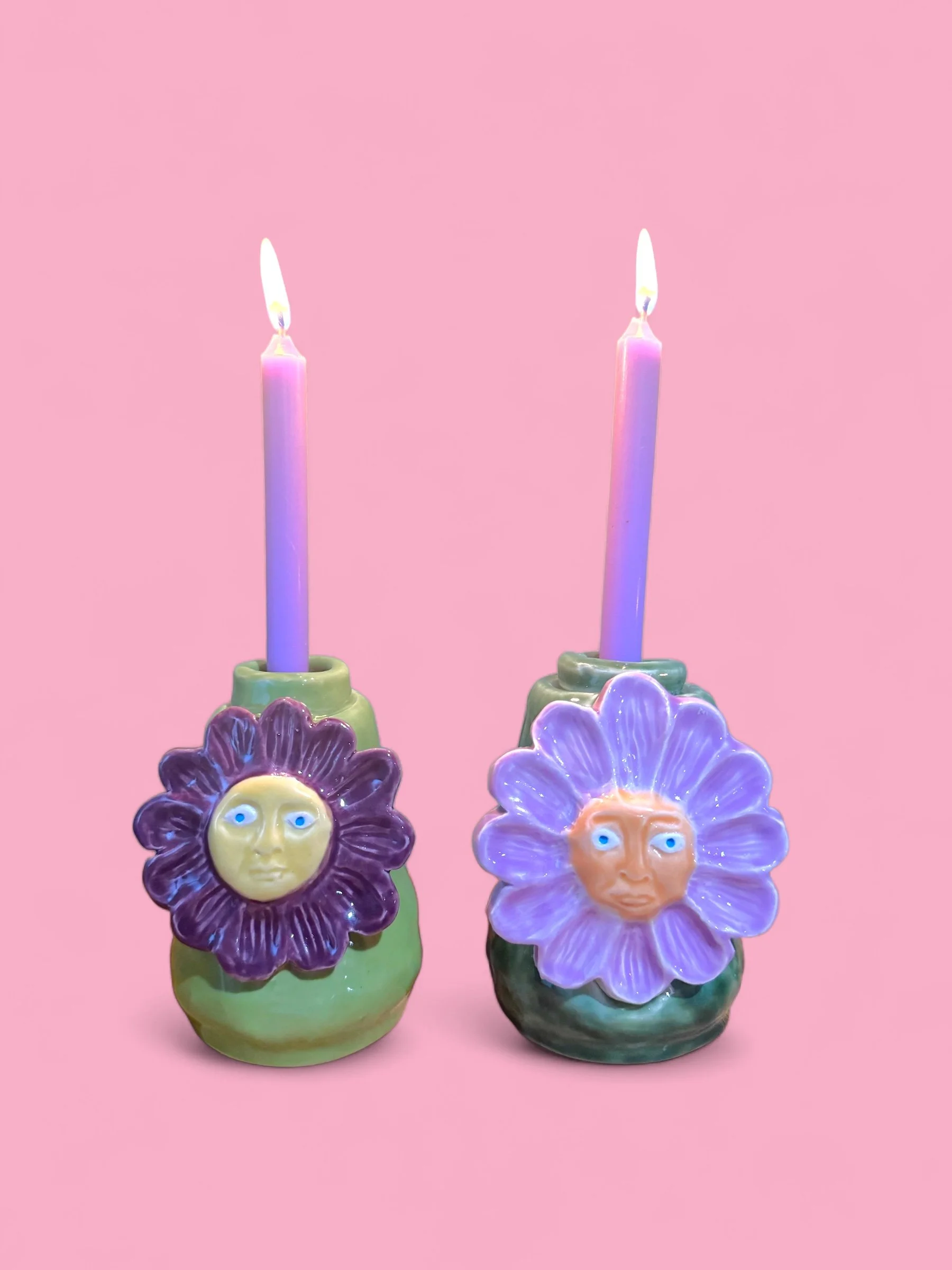 Two ceramic flower-shaped candle holders with face designs, holding lit purple candles, on a pink background.