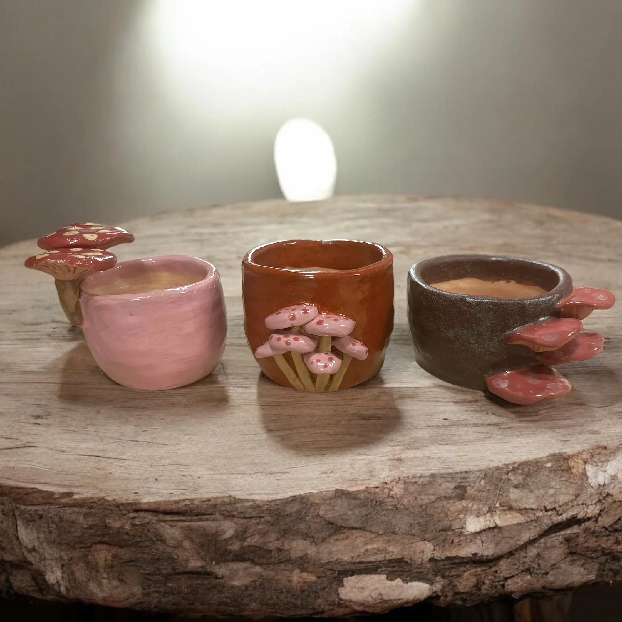 Three small handcrafted ceramic cups with mushroom and hand designs sit on a wooden log. The cups are pink, brown, and dark gray, each decorated with edible-looking mushroom shapes.