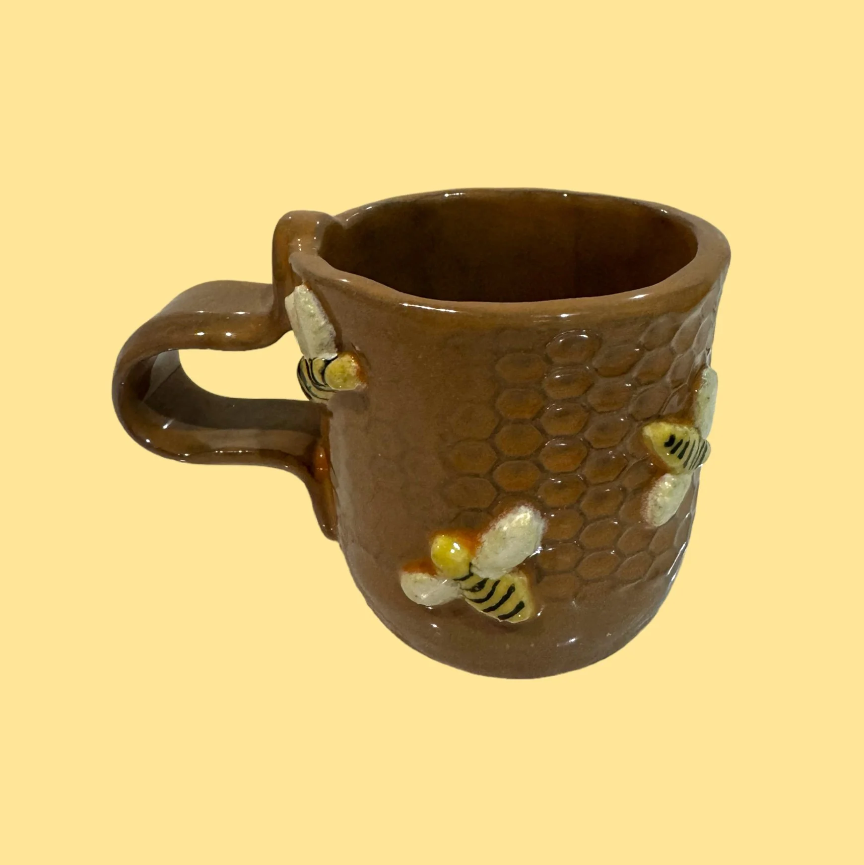 A brown ceramic mug decorated with a honeycomb pattern and small honey bee designs, set against a light yellow background.