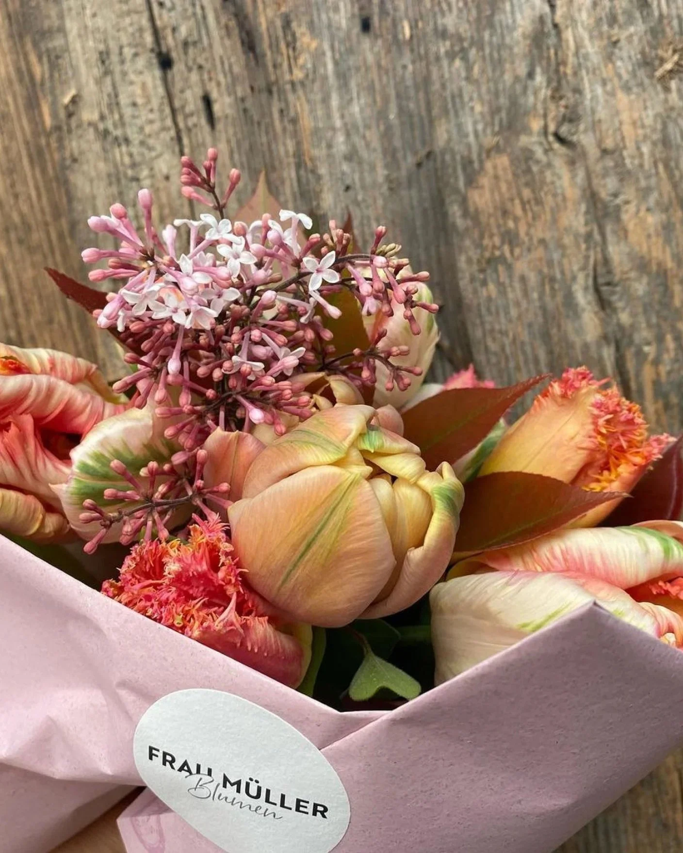 Blumenstrauß mit pinken und orangefarbenen Blumen, eingewickelt in rosa Papier, auf Holzuntergrund.