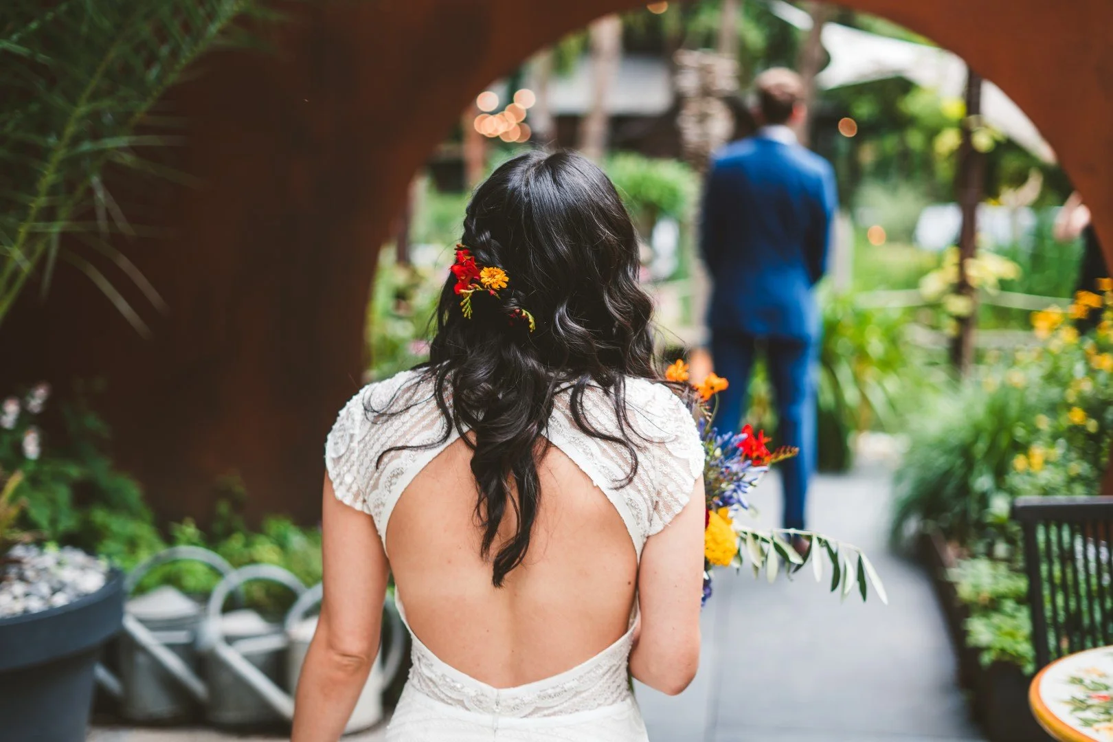Frau in weißem Hochzeitskleid mit offenem Rücken, dunkle Haare mit Blumen, Blick auf einen Mann in blauem Anzug bei einer Hochzeitszeremonie im Garten.