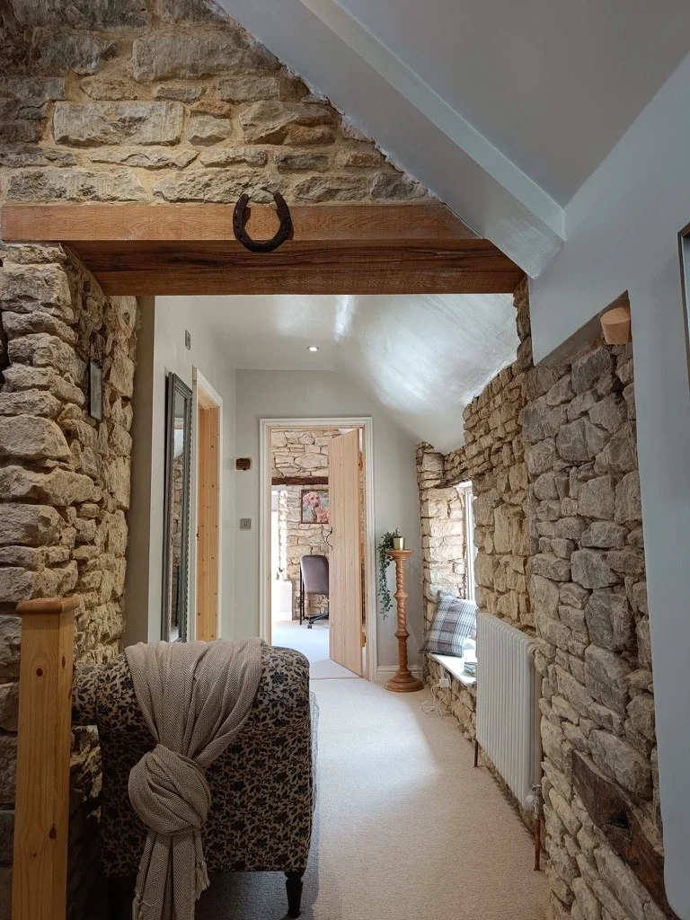 Interior view of a hallway with stone walls, a neutral-toned carpet, a wooden beam with a horseshoe hanging on it, a leopard-print armchair with a beige scarf, a tall wooden plant stand with a potted plant, and a small window with a pillow on the sil