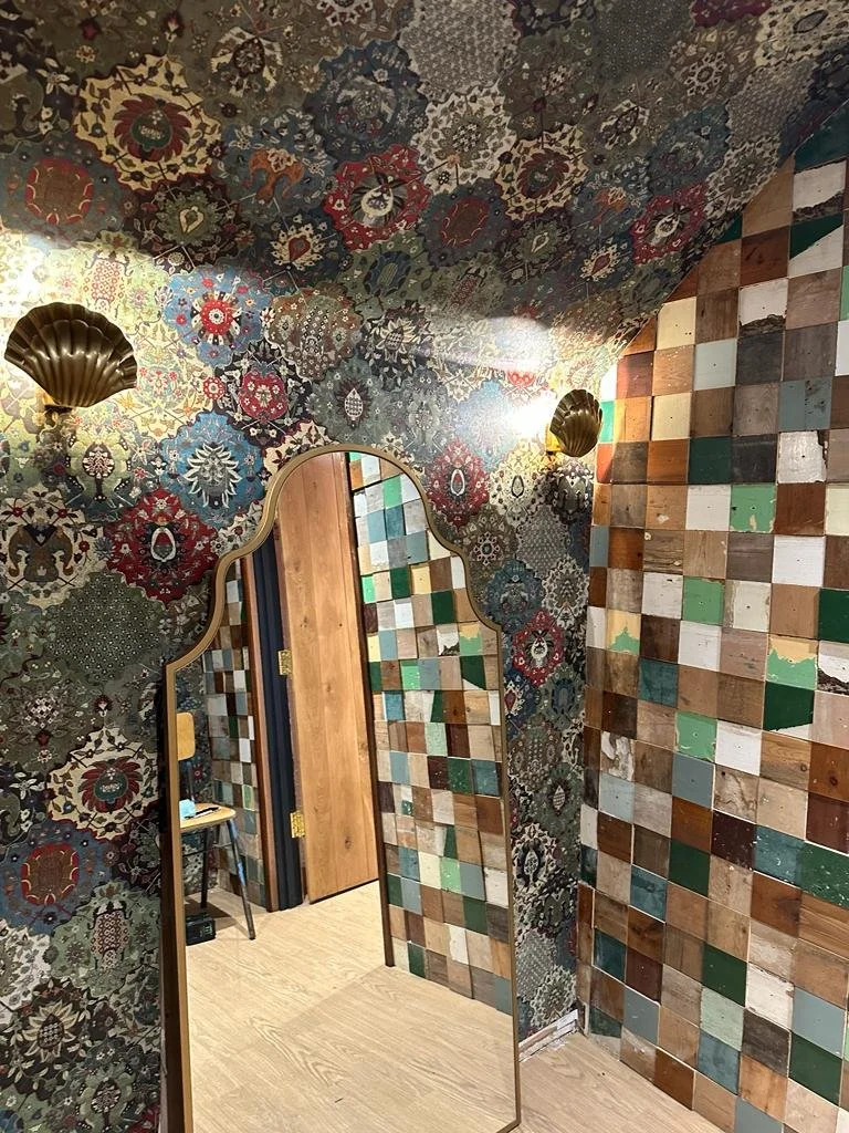 Interior room with colorful patterned wallpaper on the ceiling and a wall of mixed wooden tiles. A gold-colored decorative wall sconce and a mirror with a wavy frame reflect the room and doorway.
