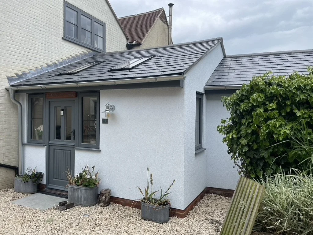 Front view of a small white house with gray window frames and door, gray slate roof, skylights, and a small gravel yard with potted plants and shrubs.