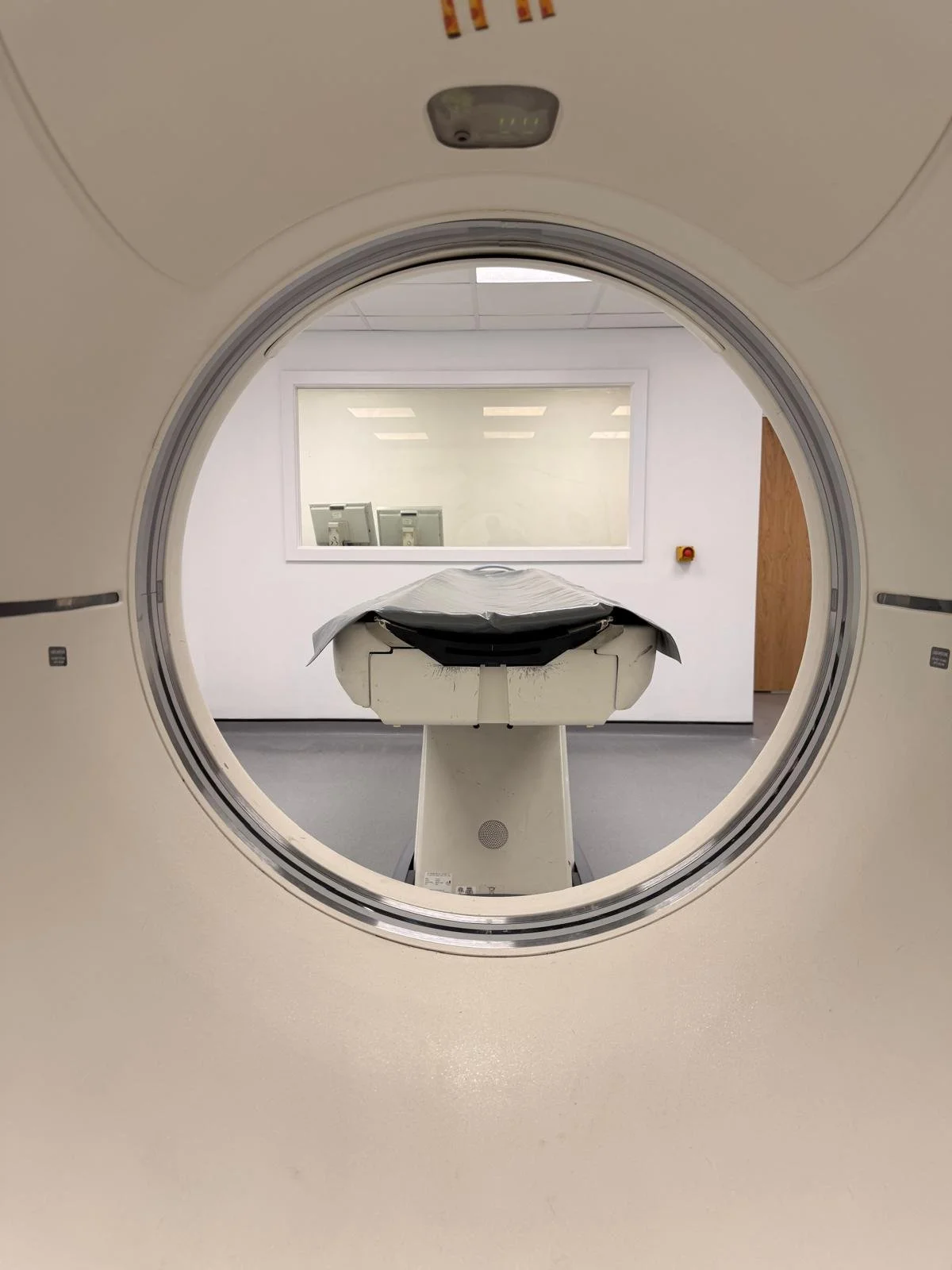 Medical imaging room with a large, empty MRI or CT scanner viewed through the circular opening, with the patient table in the center.