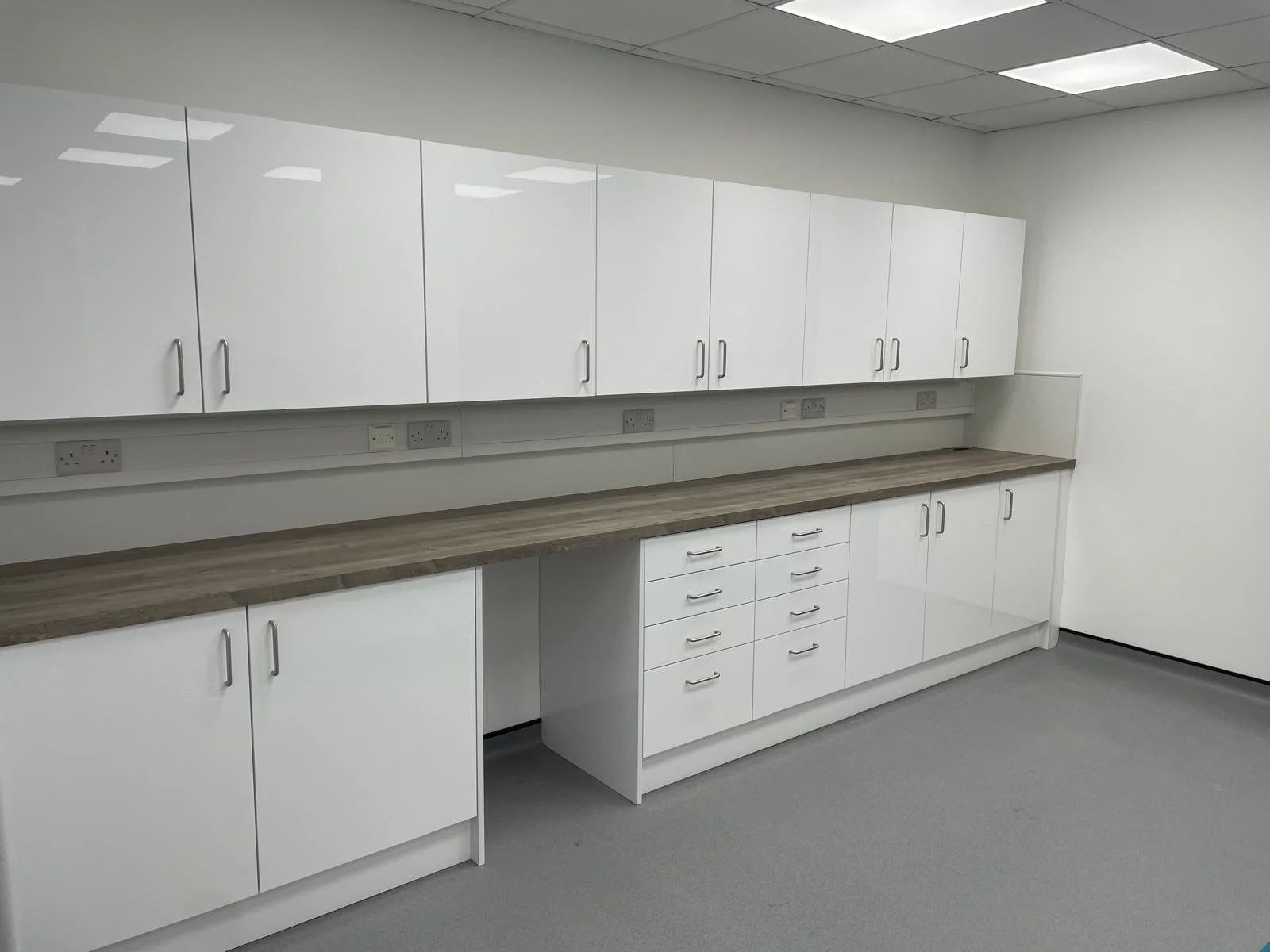 Empty modern office kitchenette with white cabinets, a woodgrain countertop, electrical outlets, and a gray carpeted floor.