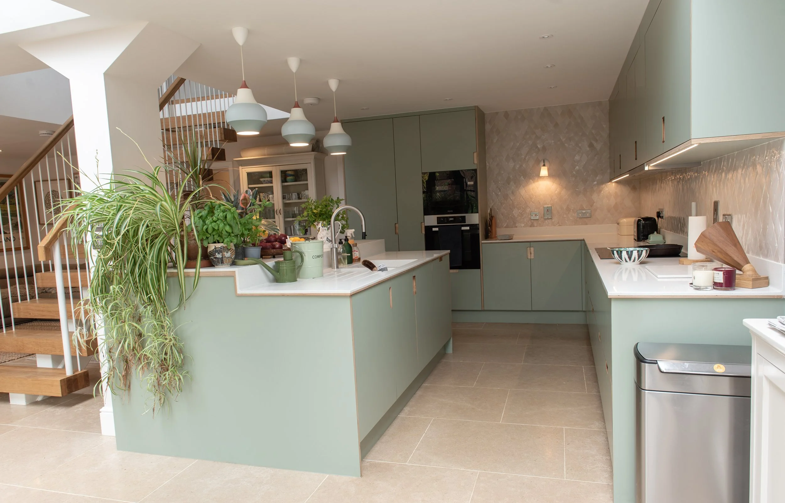 Modern kitchen with green cabinets, white countertops, hanging pendant lights, and potted plants on the island.