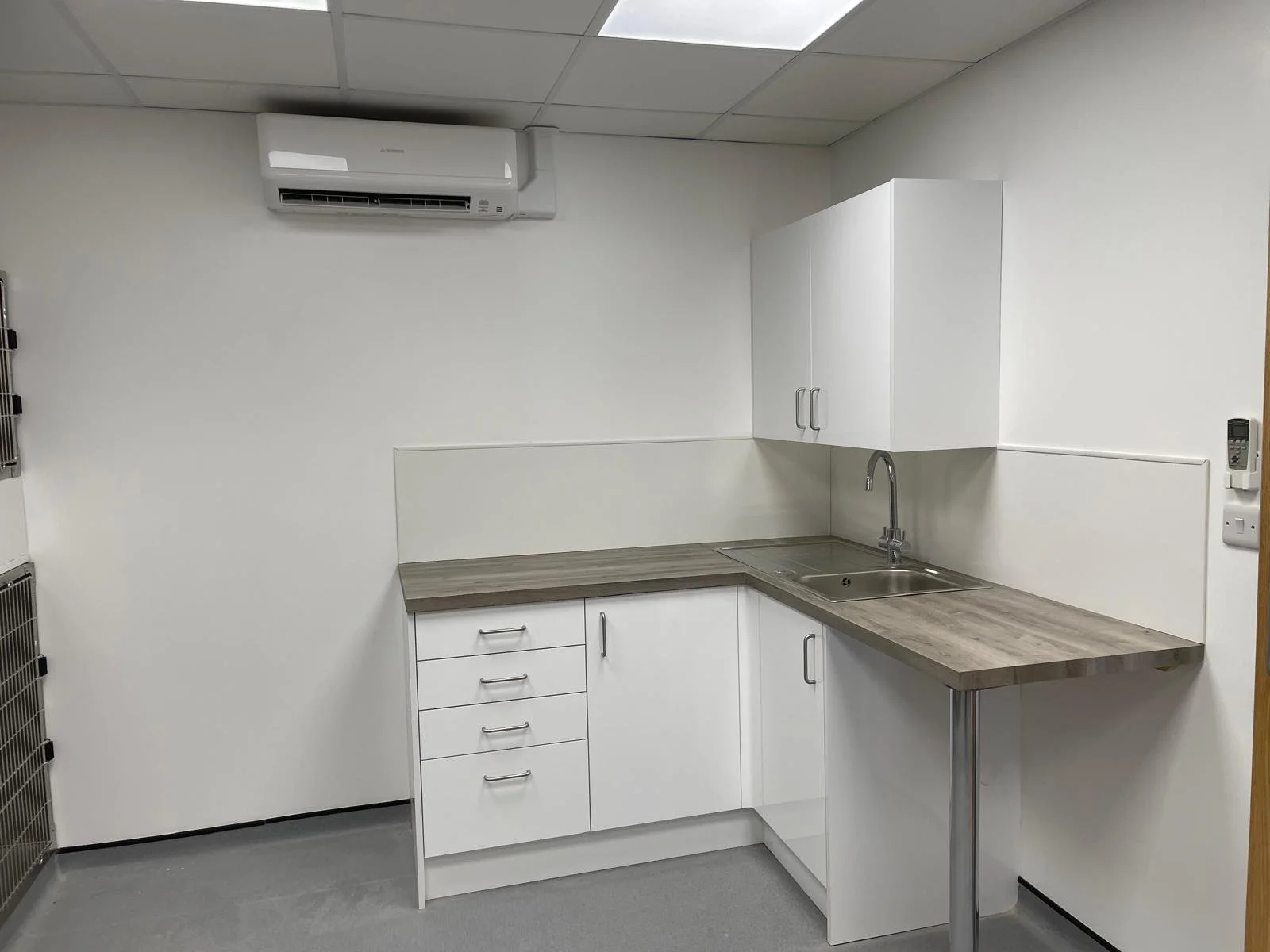 Empty kitchenette with white cabinets, gray countertop, stainless steel sink, wall-mounted air conditioning unit, and tiled ceiling in a sterile room.