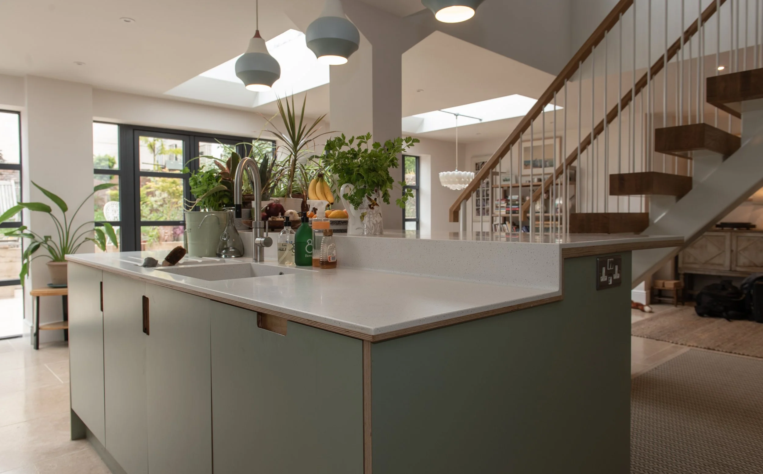 Modern kitchen with white island counter, green cabinets, and a skylight. Various plants and fruits on the countertop. Staircase with wooden steps and white railing. Large windows and open living space.