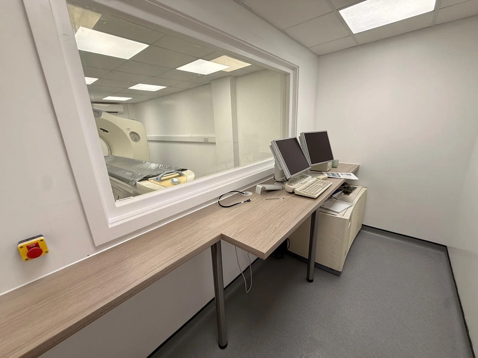 A medical exam room with a computer workstation on a wooden counter, a large window looking into an adjacent room with a medical imaging device, and a small control panel on the wall.
