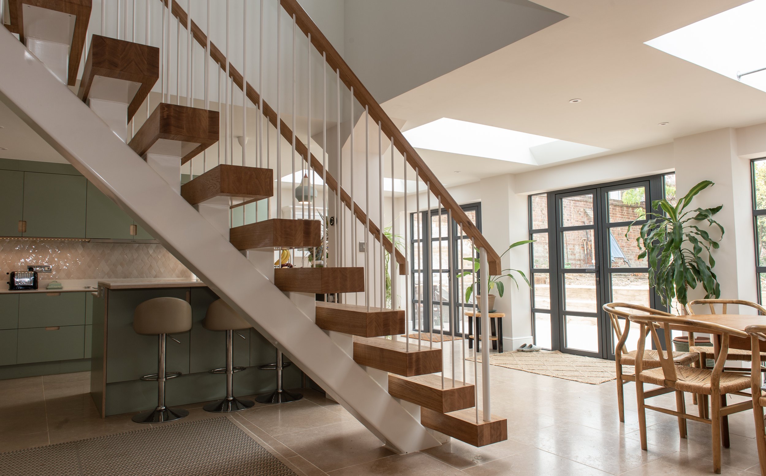 Modern interior with a staircase featuring wooden steps and white railing, large windows, green kitchen cabinets, bar stools, and a dining area with wooden chairs, natural light, and indoor plants.