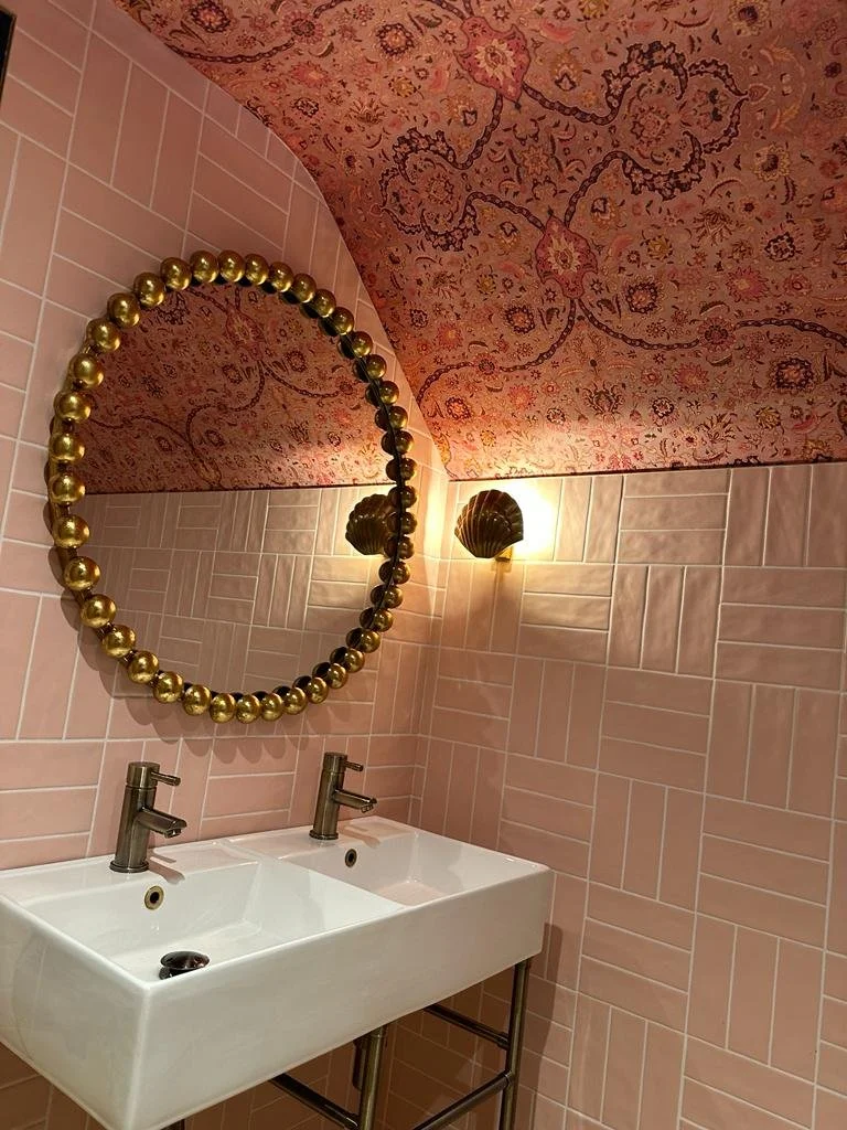 A bathroom with pink tiled walls, a pink patterned vaulted ceiling, an oval mirror decorated with gold balls, two shell-shaped wall sconces, and a white double sink with modern faucets.