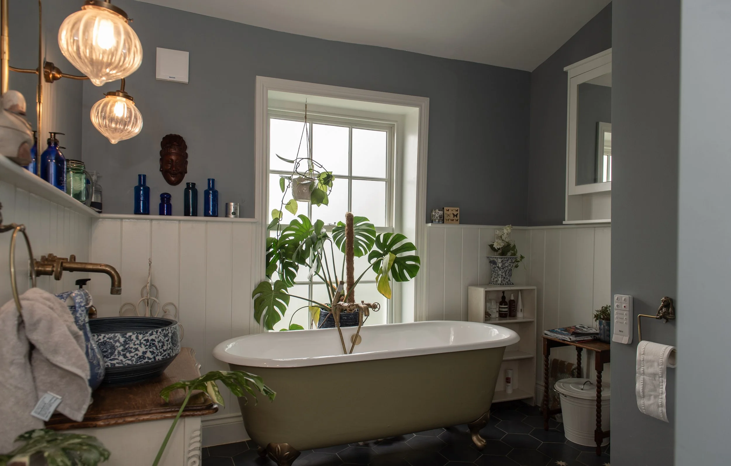 A vintage clawfoot bathtub with a large green monstera plant inside, located in a bathroom with gray walls, white beadboard paneling, and a large window with sunlight coming through.