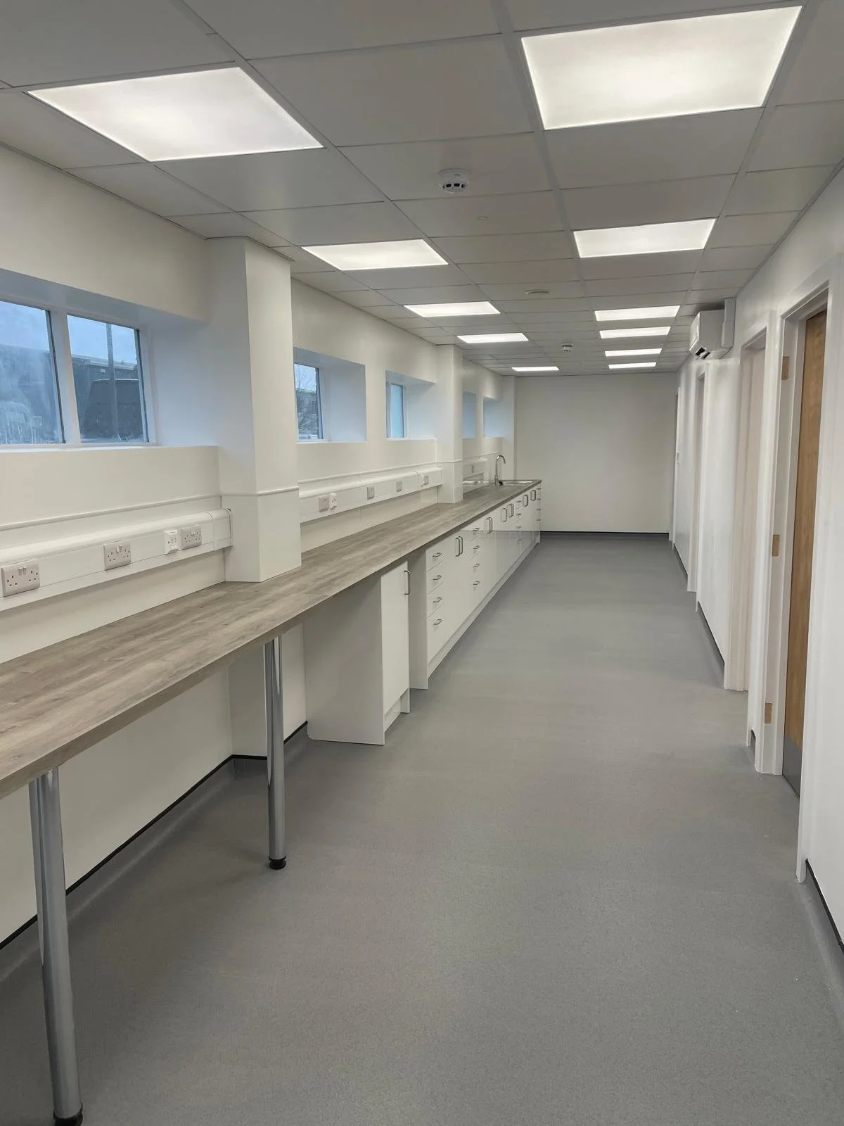 Empty laboratory or medical room with long countertop, multiple electrical outlets, windows, and closed doors.