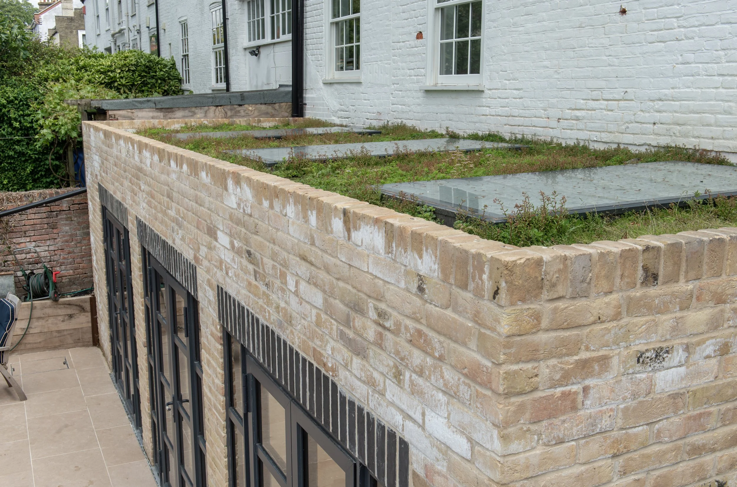 A backyard patio area with a raised brick wall, a white brick house with multiple windows, and a grassy rooftop garden with glass covers, plants, and bushes nearby.