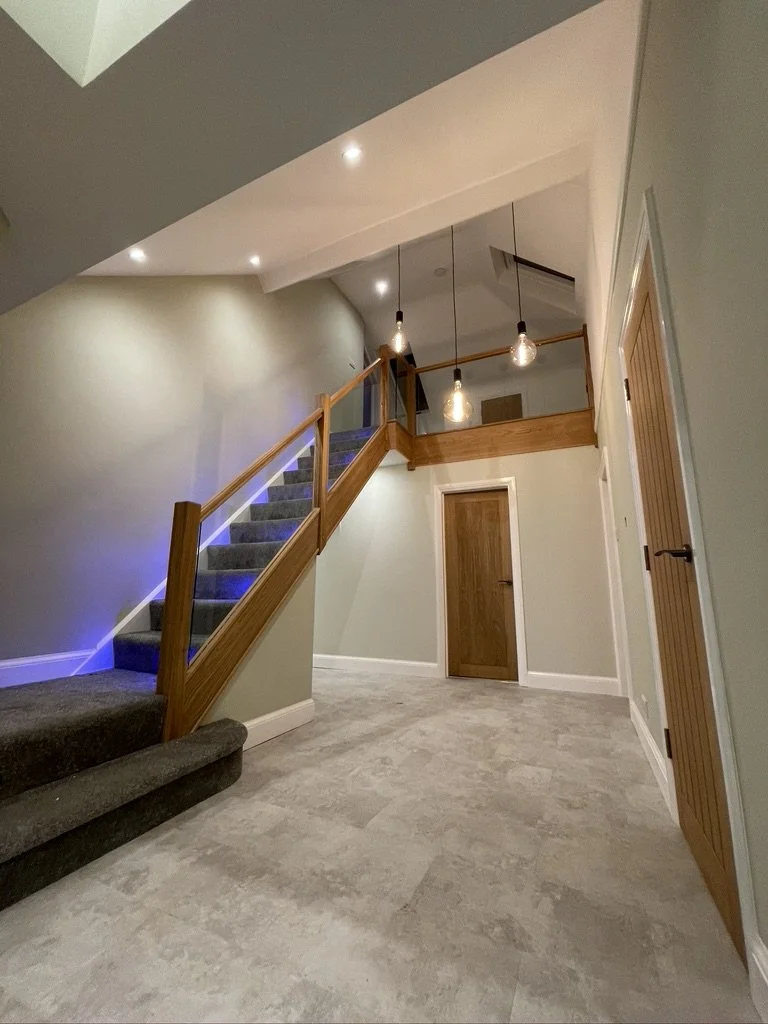 Interior view of a modern house with a staircase, wooden railings, hanging pendant lights, and light-colored walls and flooring.