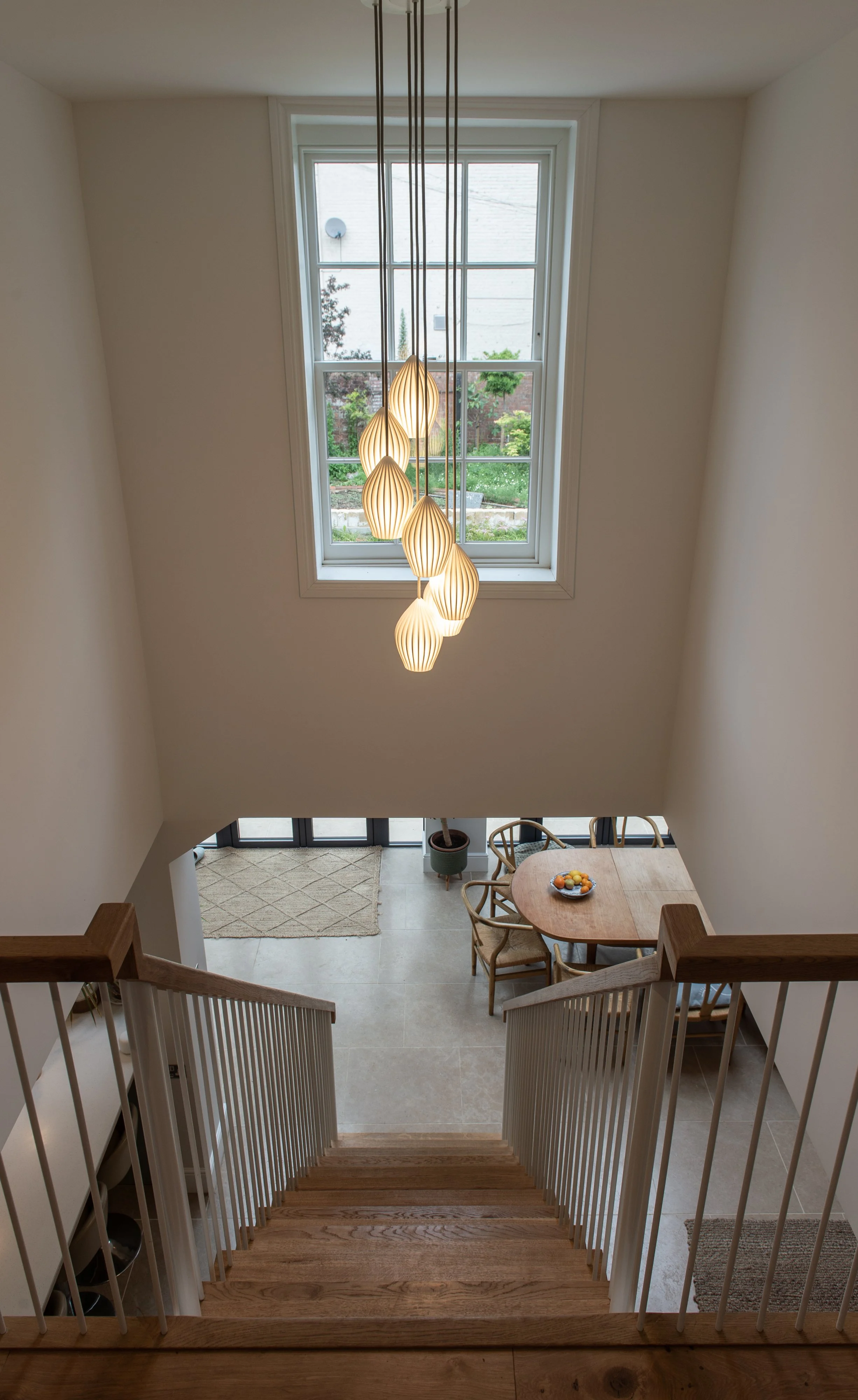 A view from the top of a staircase looking down into a bright dining area with a round wooden table, chairs, a fruit bowl, a large window, and a modern hanging light fixture.