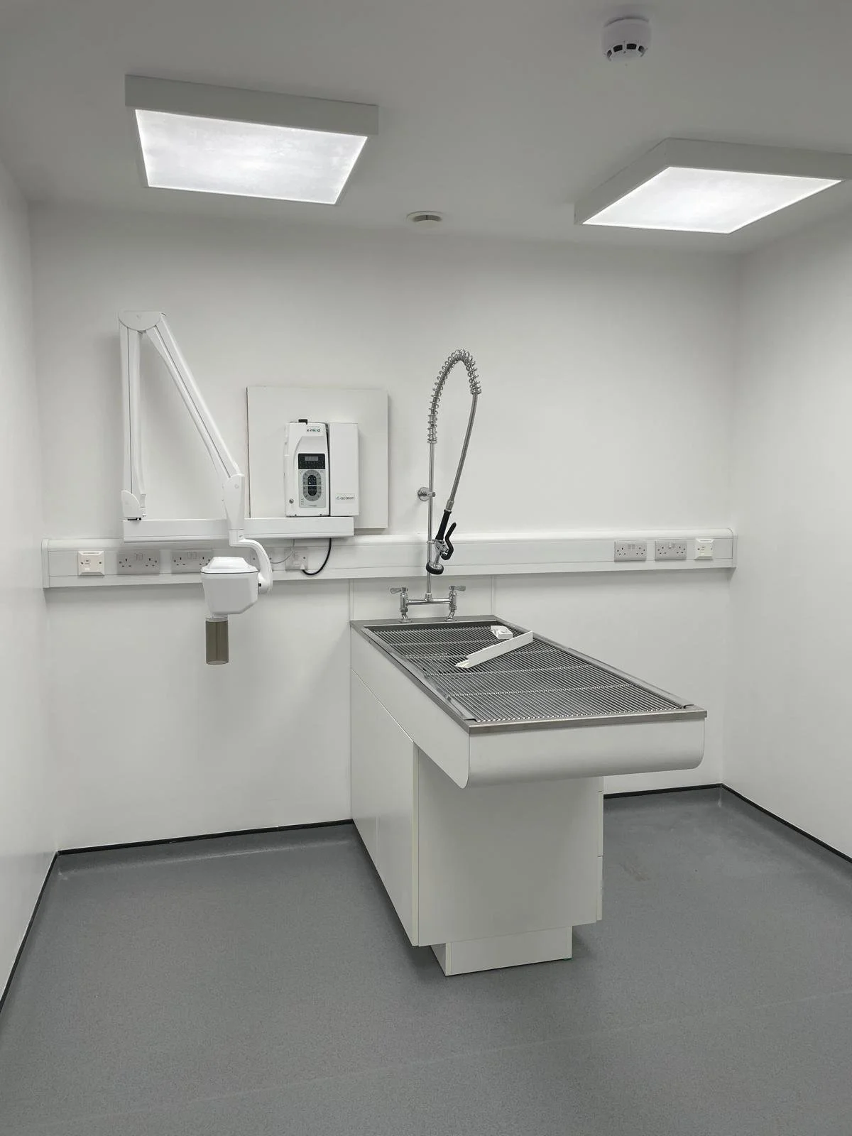 A clean, modern veterinary examination room with a stainless steel sink, an adjustable faucet, and medical equipment mounted on the white wall, including a sterilizer and electrical outlets. The room has bright overhead lighting and a grey floor.