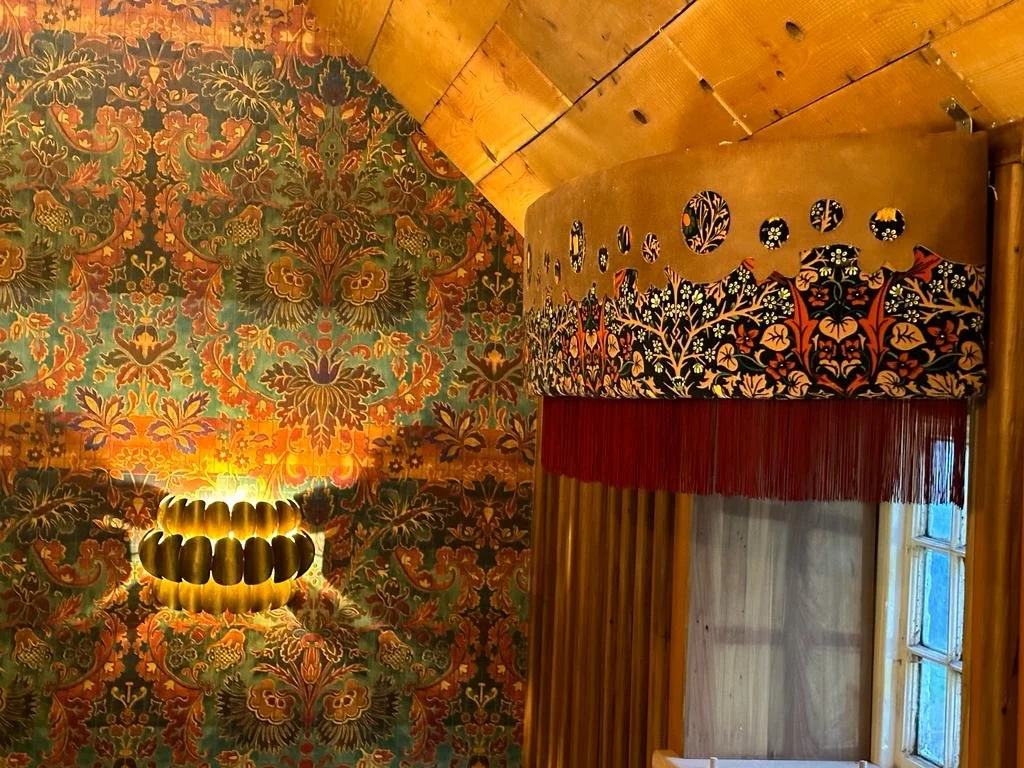 Decorative interior featuring a patterned wall with floral motifs, a wall sconce with a gold-colored design, a wooden window and an ornate valance with floral embroidery and red fringe.