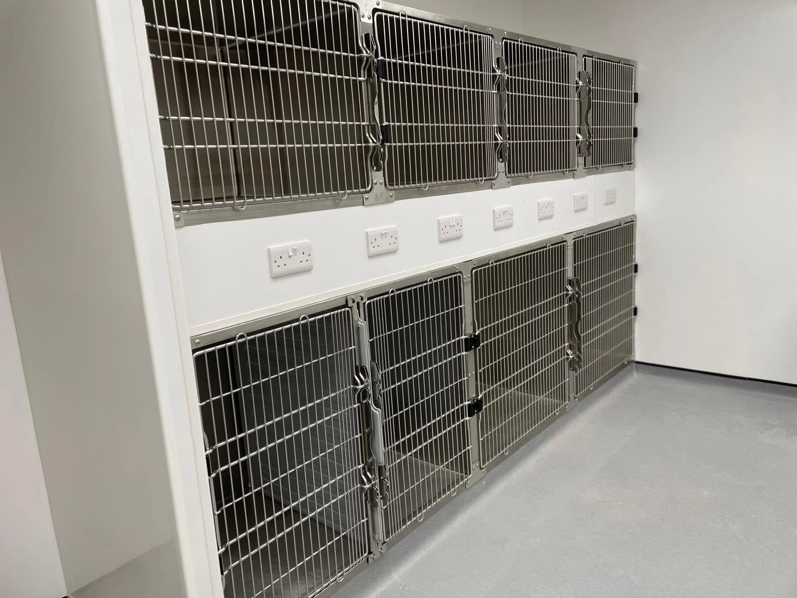 A row of stainless steel animal cages or kennels mounted on a white wall, with electrical outlets beneath them, in a clean and sterile environment.