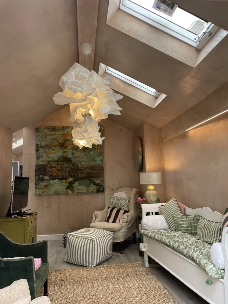 Living room with beige walls, a unique cloud-shaped pendant light, a large abstract painting, a beige armchair with pillows, a green armchair, a white daybed with pillows, a small white side table with a large vase and pink flowers, a lamp, a flat-sc
