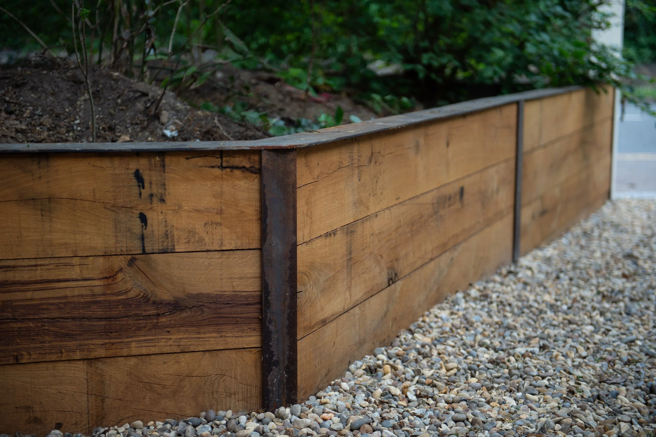 A wooden garden bed with soil on top, surrounded by gravel, with greenery in the background.