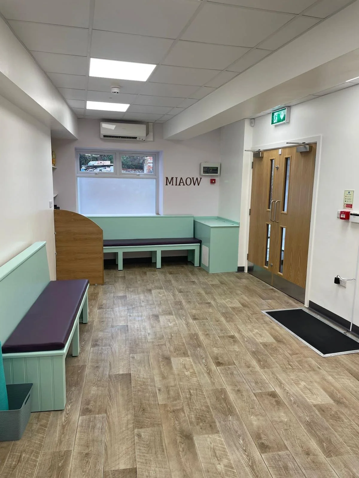 Empty reception area with benches, wood and pastel-colored furniture, a window, and a wooden door with an emergency exit sign above. The room has a wood pattern floor and ceiling with fluorescent lights.