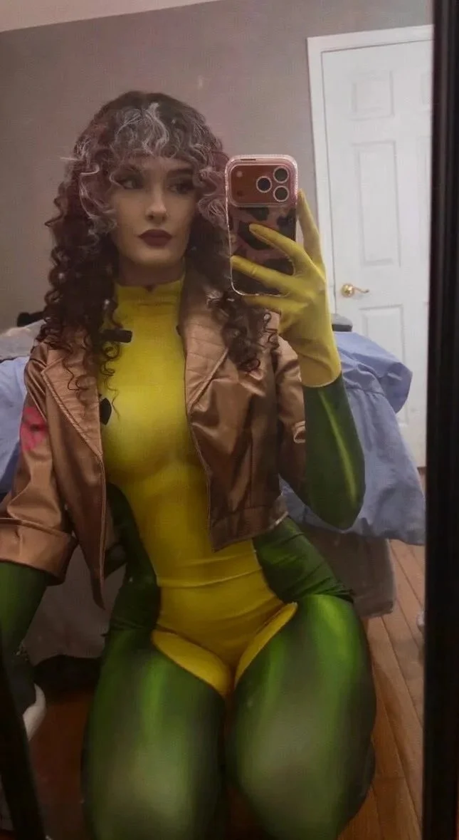 kaeleeetv taking a mirror selfie, wearing a yellow rogue from x-men cosplay bodysuit with green accents, a brown cropped jacket, and yellow gloves, with curly hair and dark lipstick.