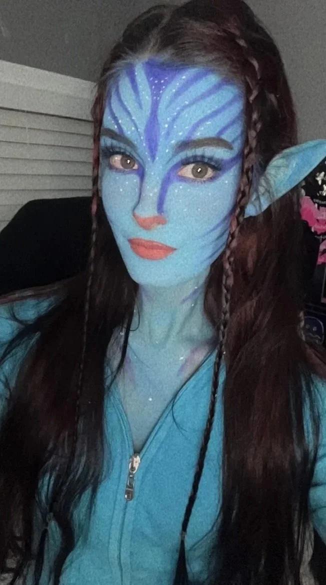 kaeleeetv with blue face and body paint resembling a avatar character, wearing a blue hoodie, with long brown hair styled in braids, and elf-like ear prosthetics.