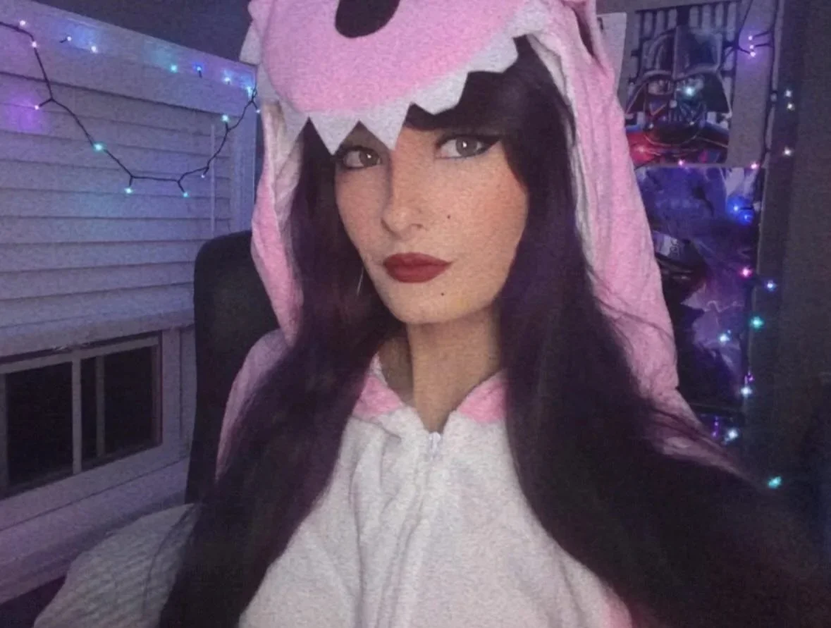 kaeleeetv with long dark hair wearing a pink and white dinosaur onesie with a hood that has a dinosaur face, sitting in a room decorated with colorful string lights and Star Wars posters in the background.