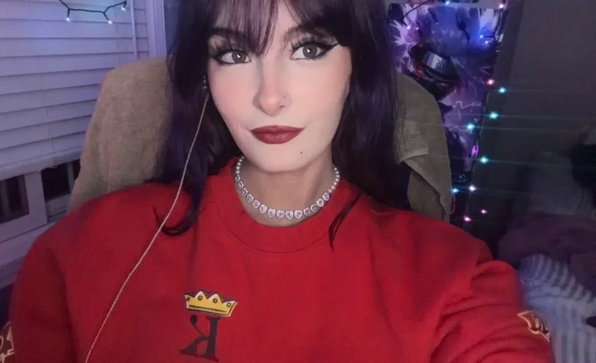 Kaeleeetv with dark hair, red lipstick, and a nose piercing, wearing a red Kaeleeetv sweatshirt with a heart-shaped diamond necklace, sitting on a beige chair with purple and blue fairy lights in the background.