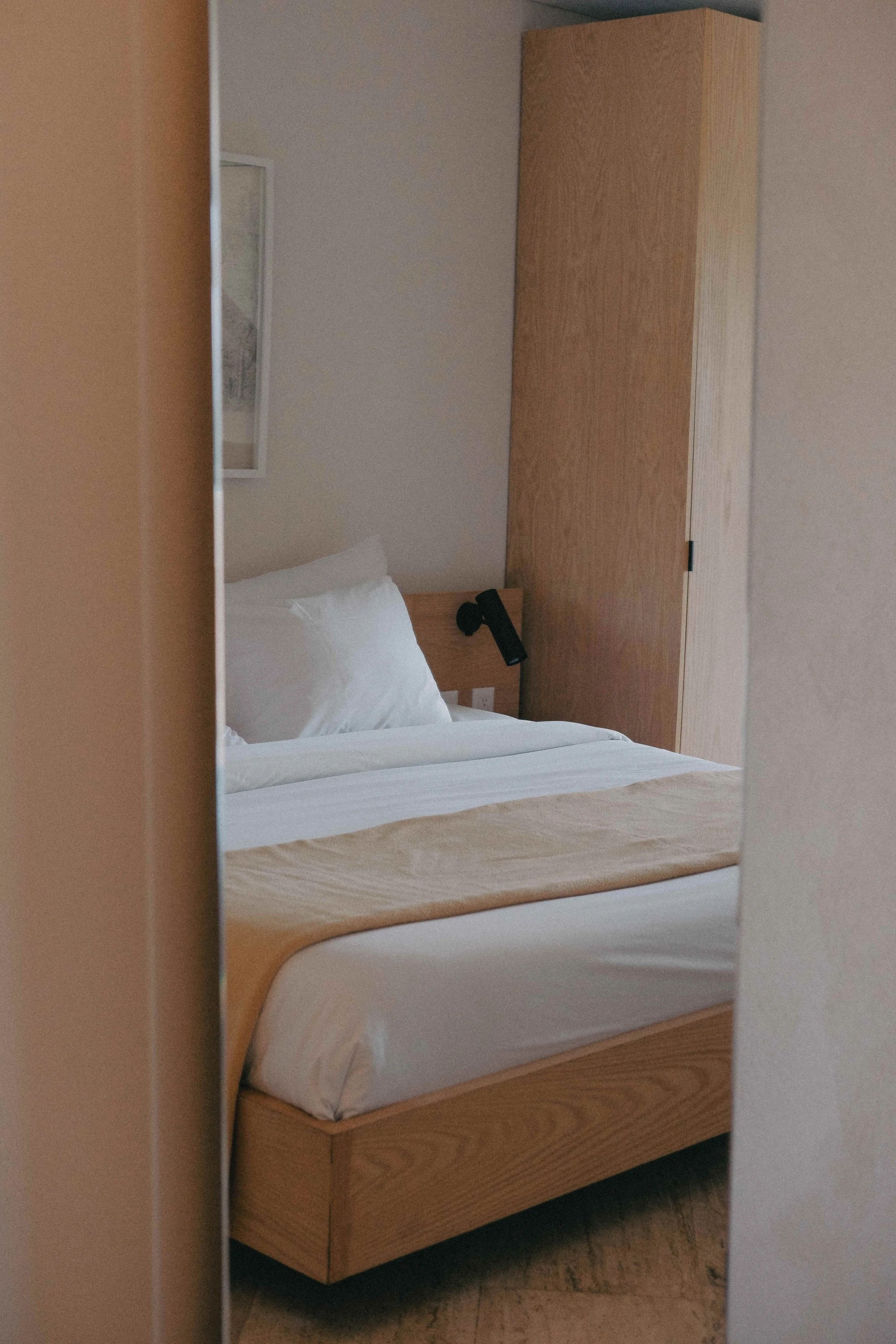 Minimalist bedroom with a made bed, tan blanket, wooden headboard, wall lamp, wooden wardrobe, and a framed picture on the wall.