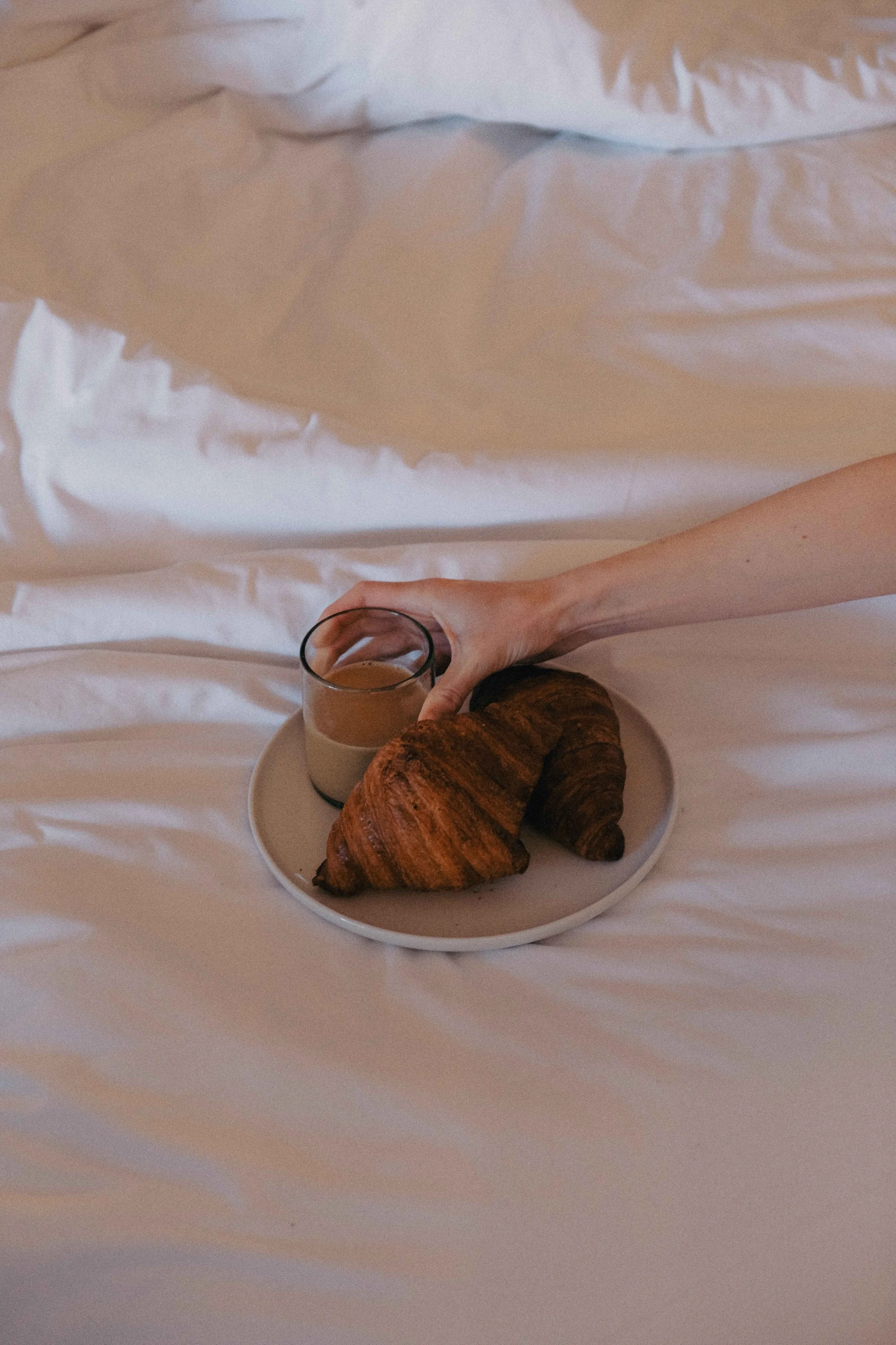A person's hand reaching for a cup of coffee next to croissants on a plate, placed on a bed with white linens.