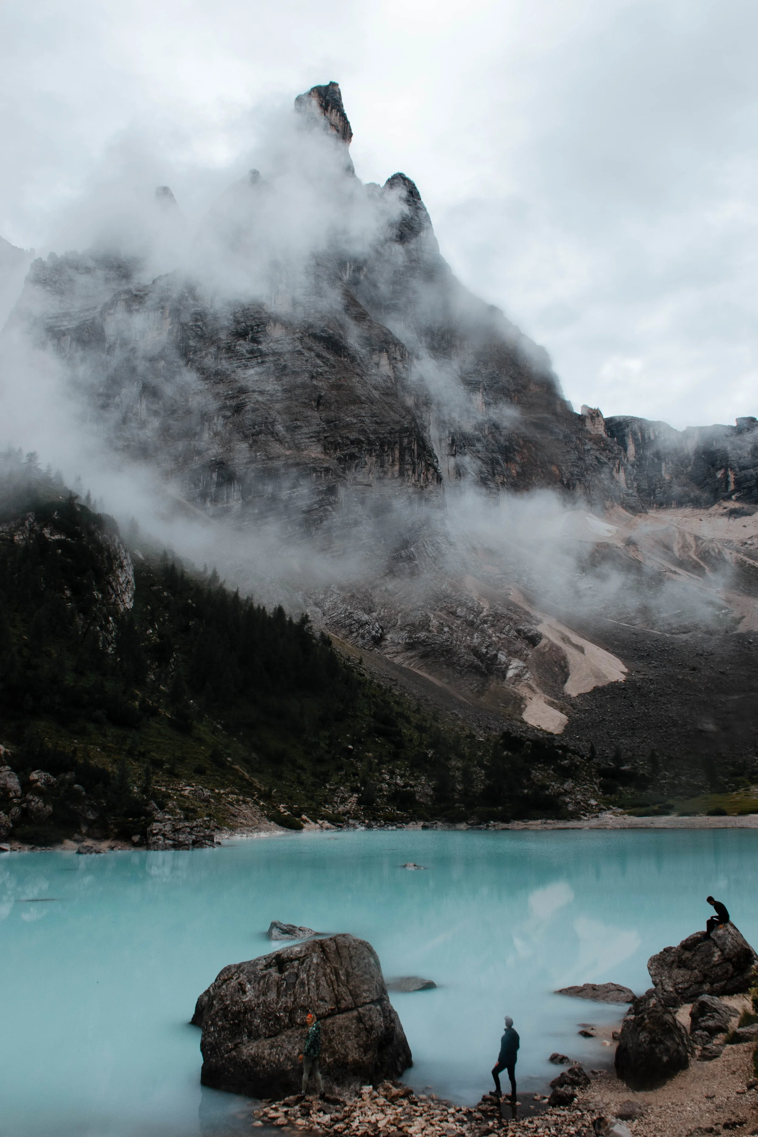 hiker standing in front of a lake and a hiker sitting on a rock in front of that same lake with tall mountain peak loomin above it