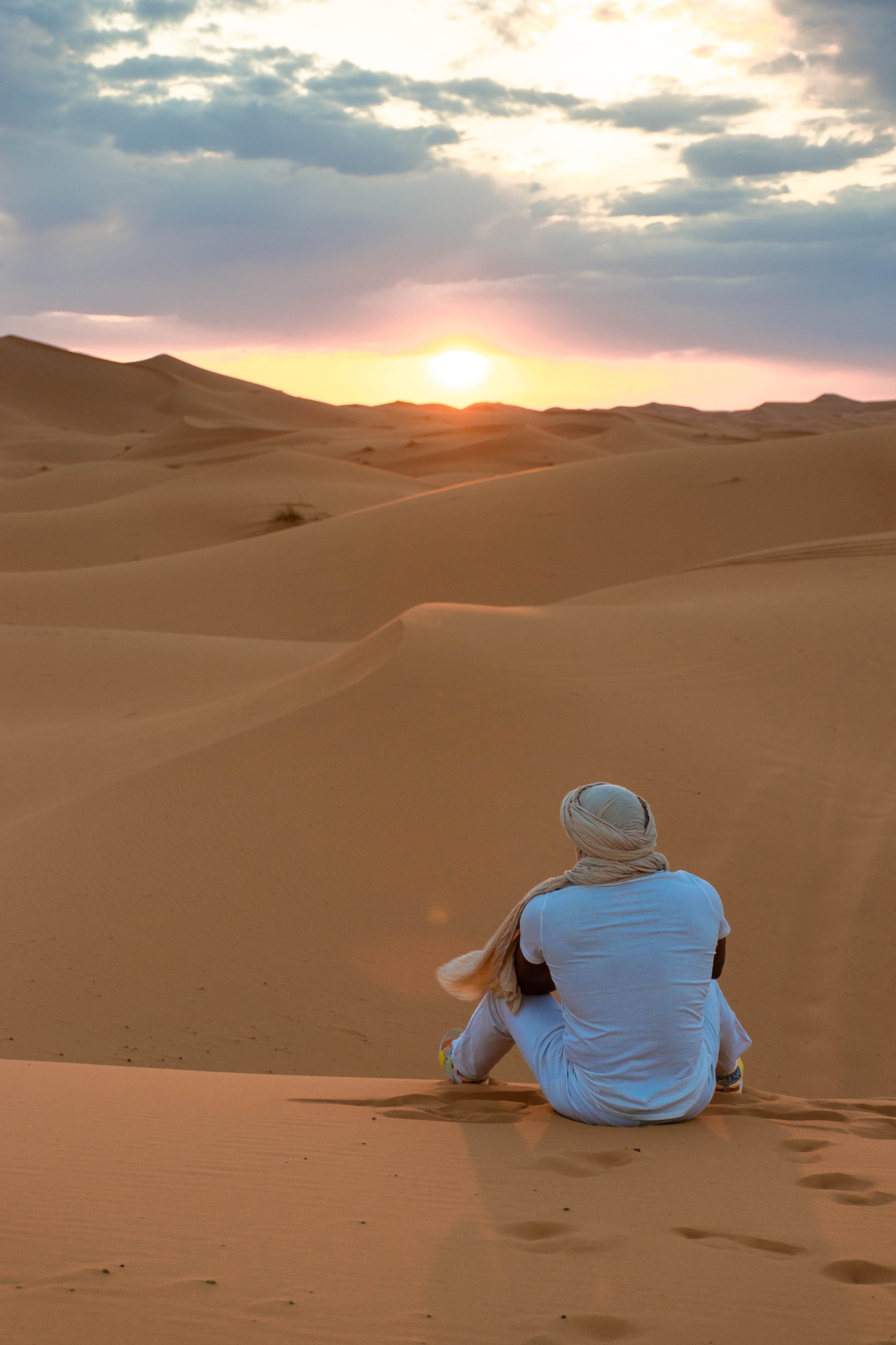 a man in white sitting on sand dunes in the desert watching the sun go below the horizon