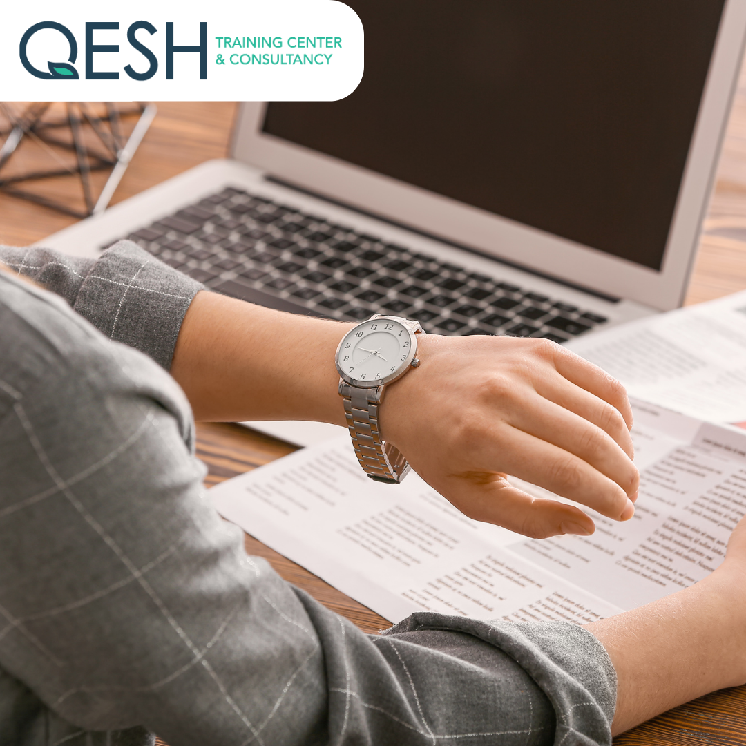 Person wearing a watch, checking time while working at a laptop with documents, QESH Training Center & Consultancy logo.