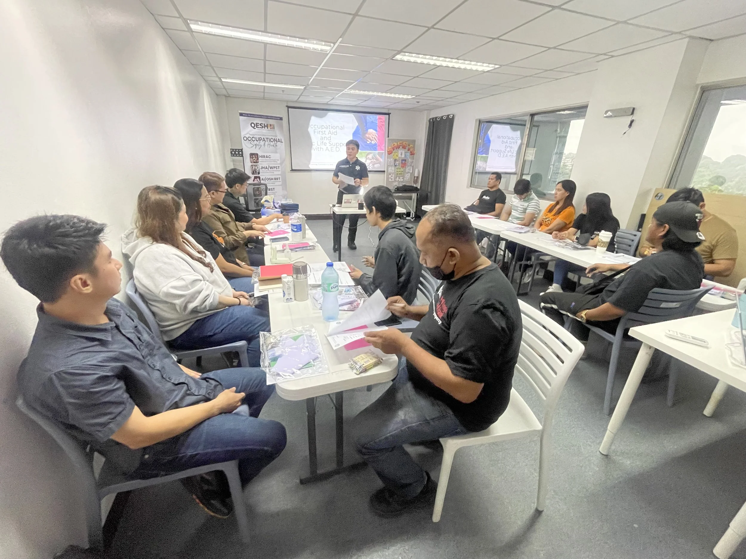 A group of people sitting around tables in a classroom setting listening to an instructor. There are presentation slides displayed in the background related to occupational first aid and life support. The room has windows and fluorescent lighting. Participants have notebooks and bottled water on the tables.