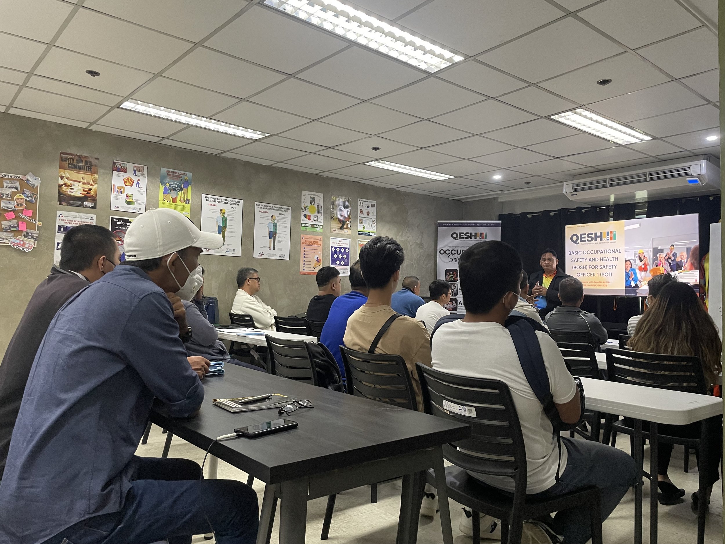 People attending a safety training session in a classroom with posters on the wall, one banner reads 'QESH Basic Occupational Safety and Health for Safety Officer 1 (SO1)'