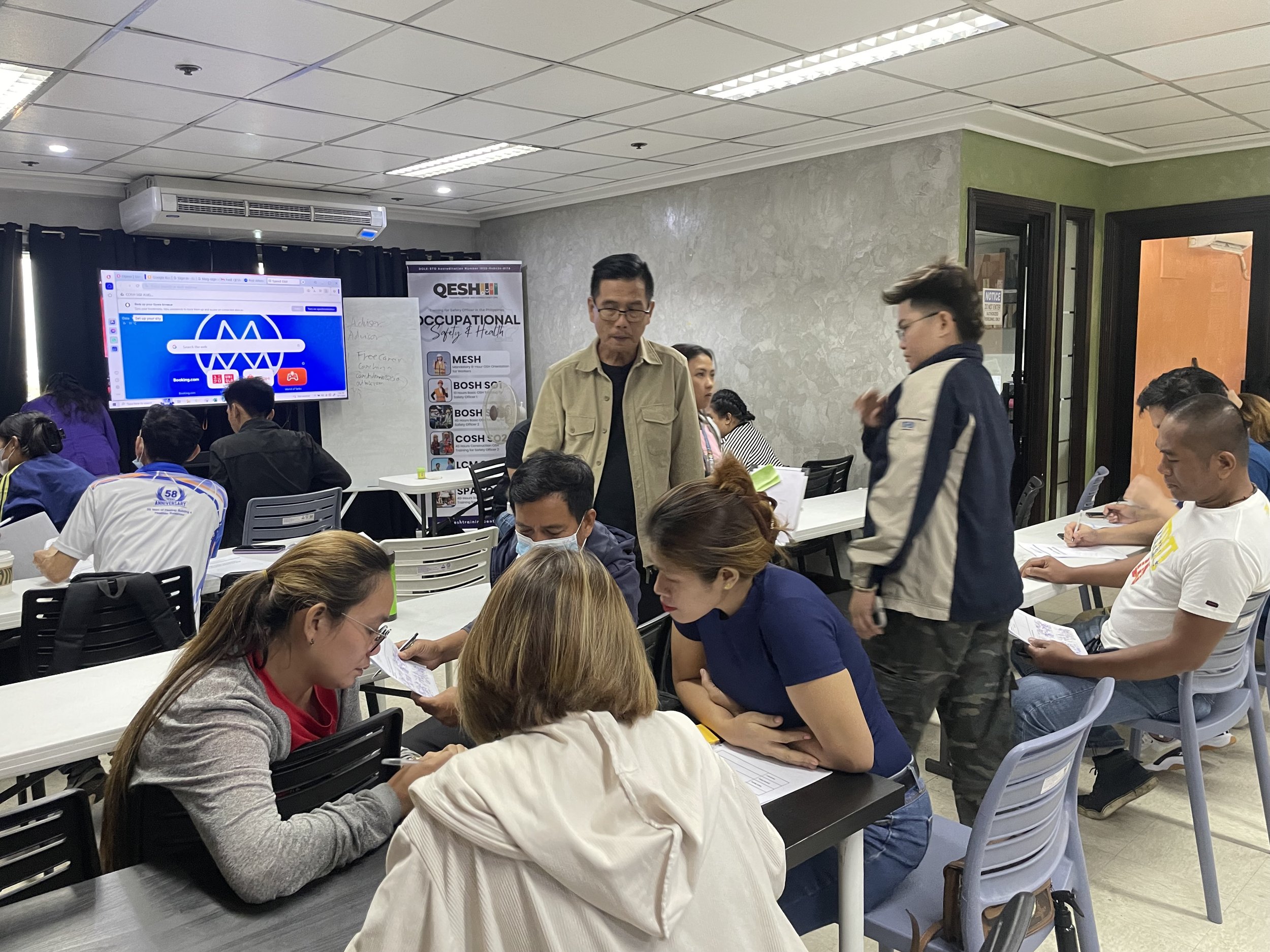 Classroom with group of people attending a training session. Participants are seated at tables, writing and discussing. A large screen displays a safety training website, and posters on occupational safety and health are visible. The room has fluorescent lighting and a modern office setting.