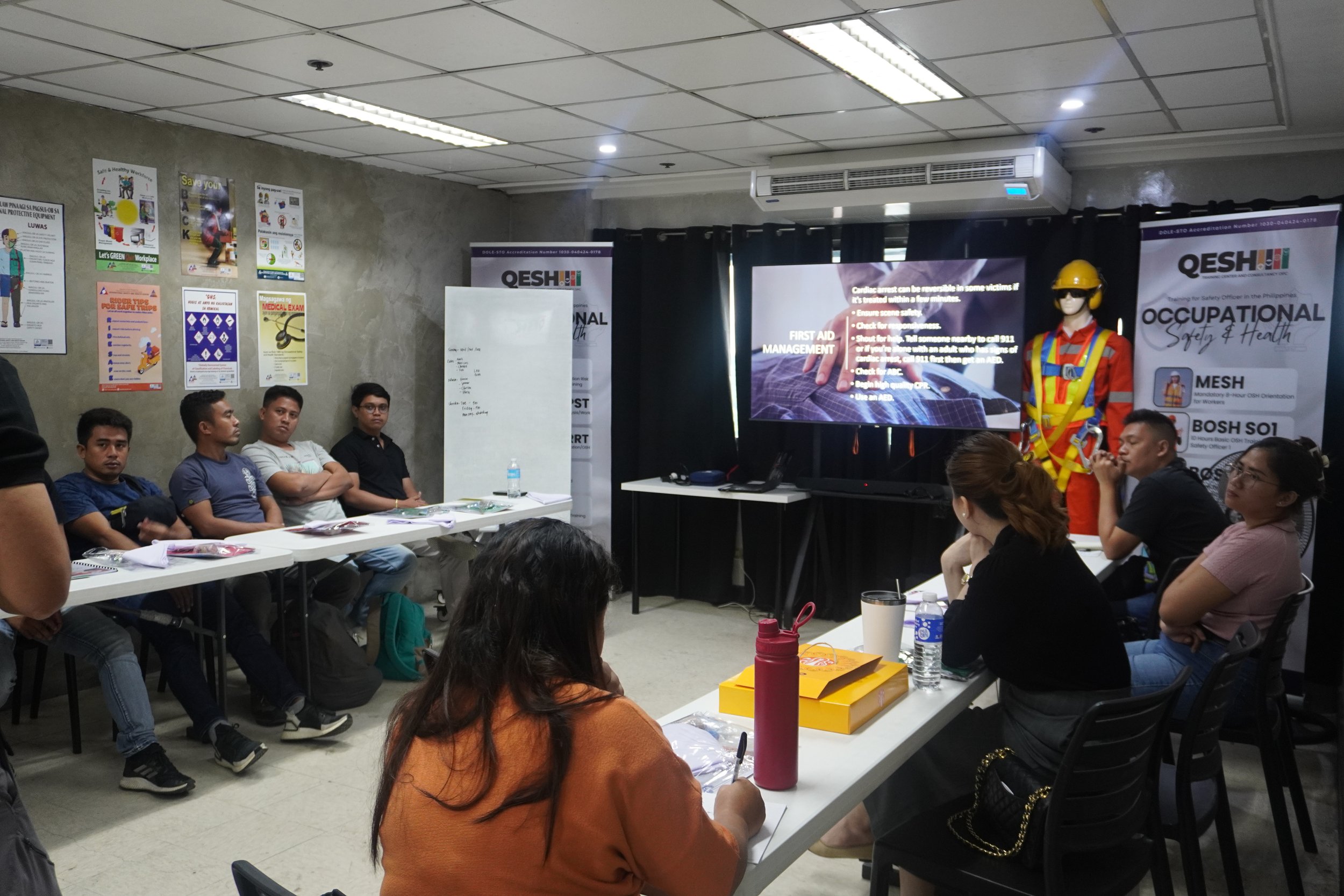 A group of people attending a safety training session in a classroom, with posters on the wall and a mannequin in safety gear beside a presentation screen displaying first aid management information.