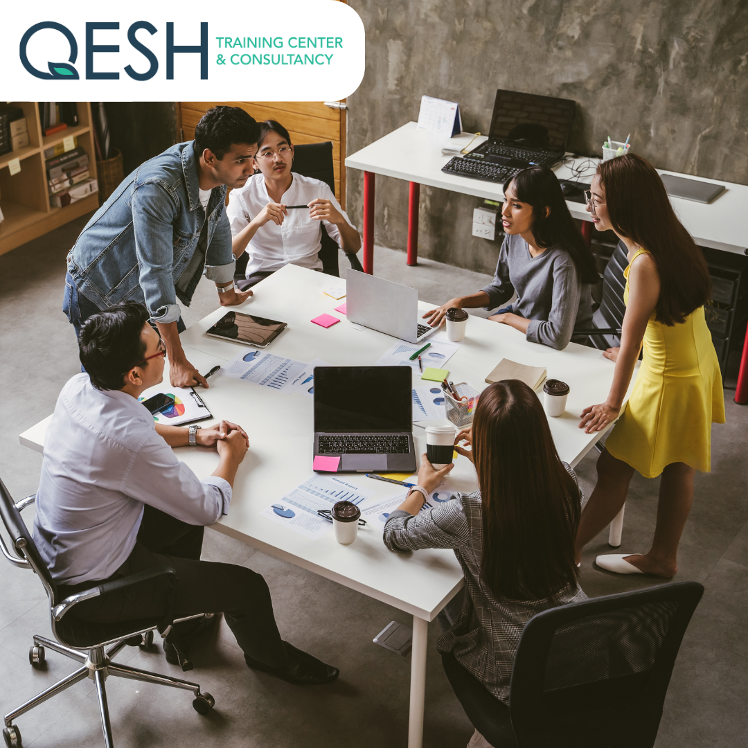 Group of people having a meeting around a table in an office setting with laptops and documents. Logo of QESH Training Center & Consultancy is visible.
