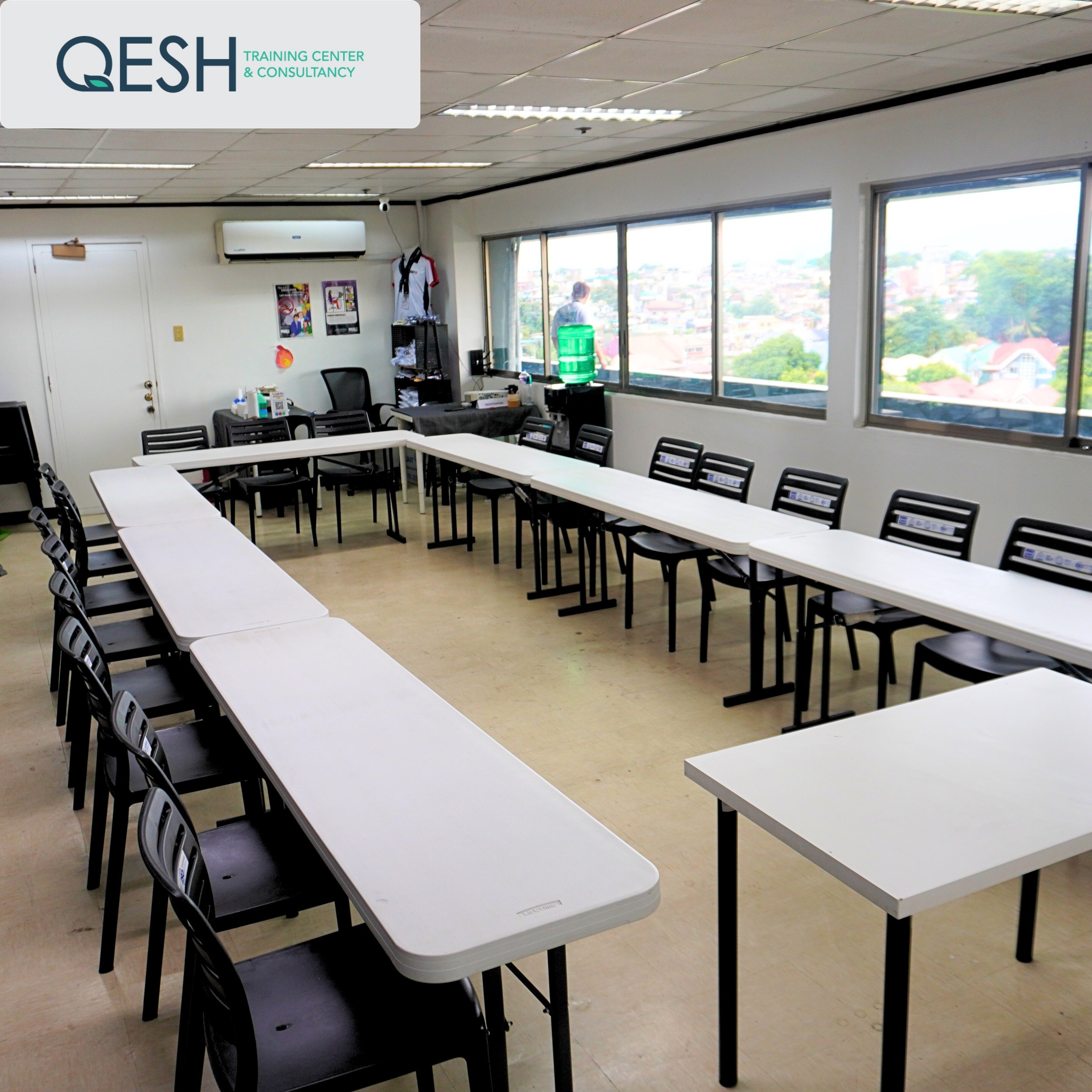 Training room with U-shaped white tables and chairs, large windows showing outside view, and wall-mounted air conditioner.