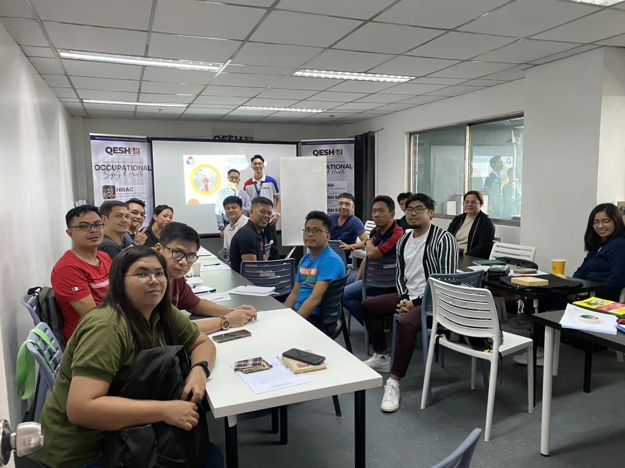 A group of people sitting around tables in a classroom setting, attending an occupational safety and health training session. A presentation is being shown on a screen at the front of the room. The room is well-lit and has several posters related to safety and health on the walls.
