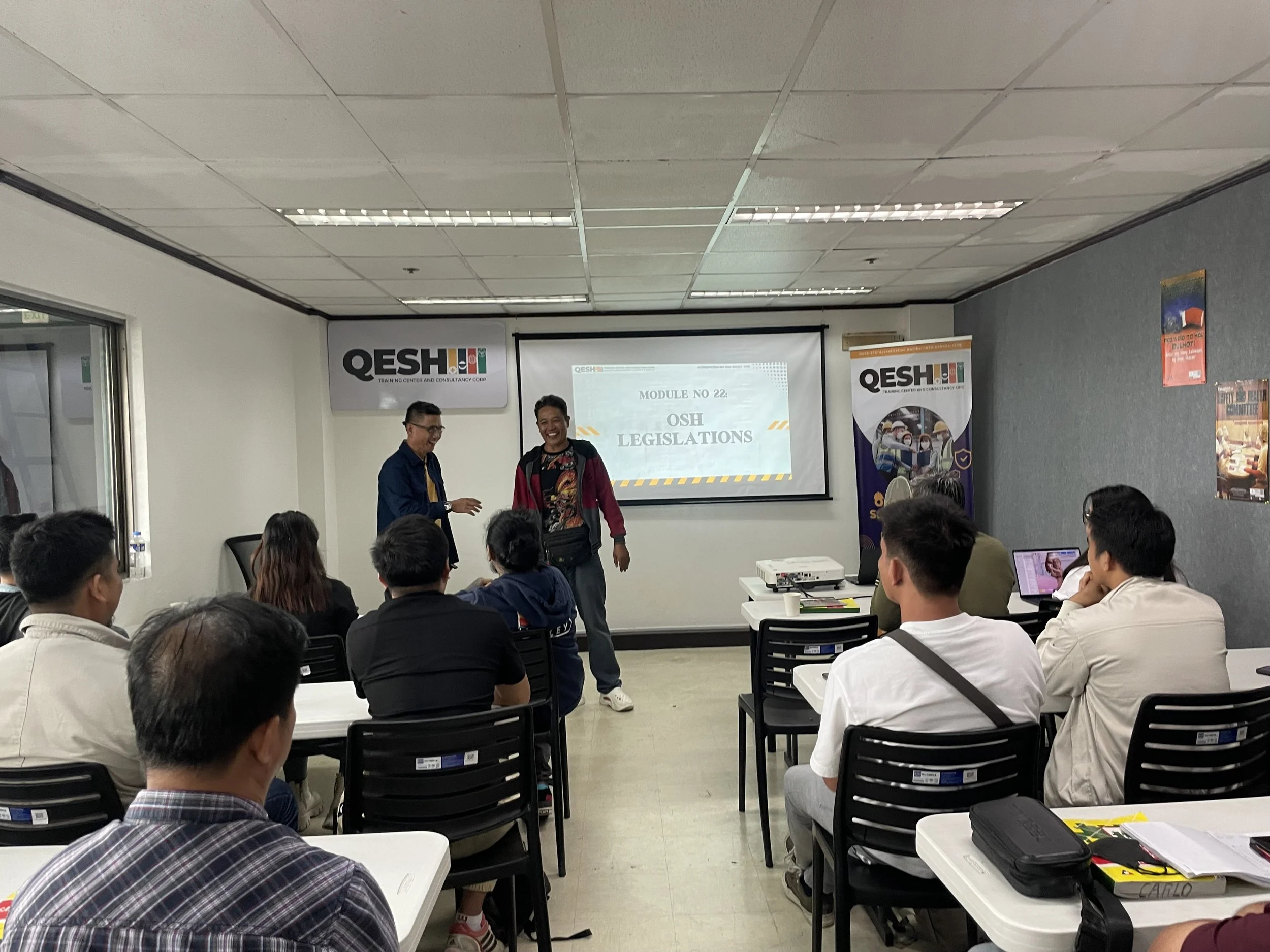 A classroom setting with people attending a session on "OSH Legislation" presented on a screen at the front. Two presenters stand near the screen, while the audience sits at desks facing forward. The room has a banner with the name "QESH" and various posters on the walls.