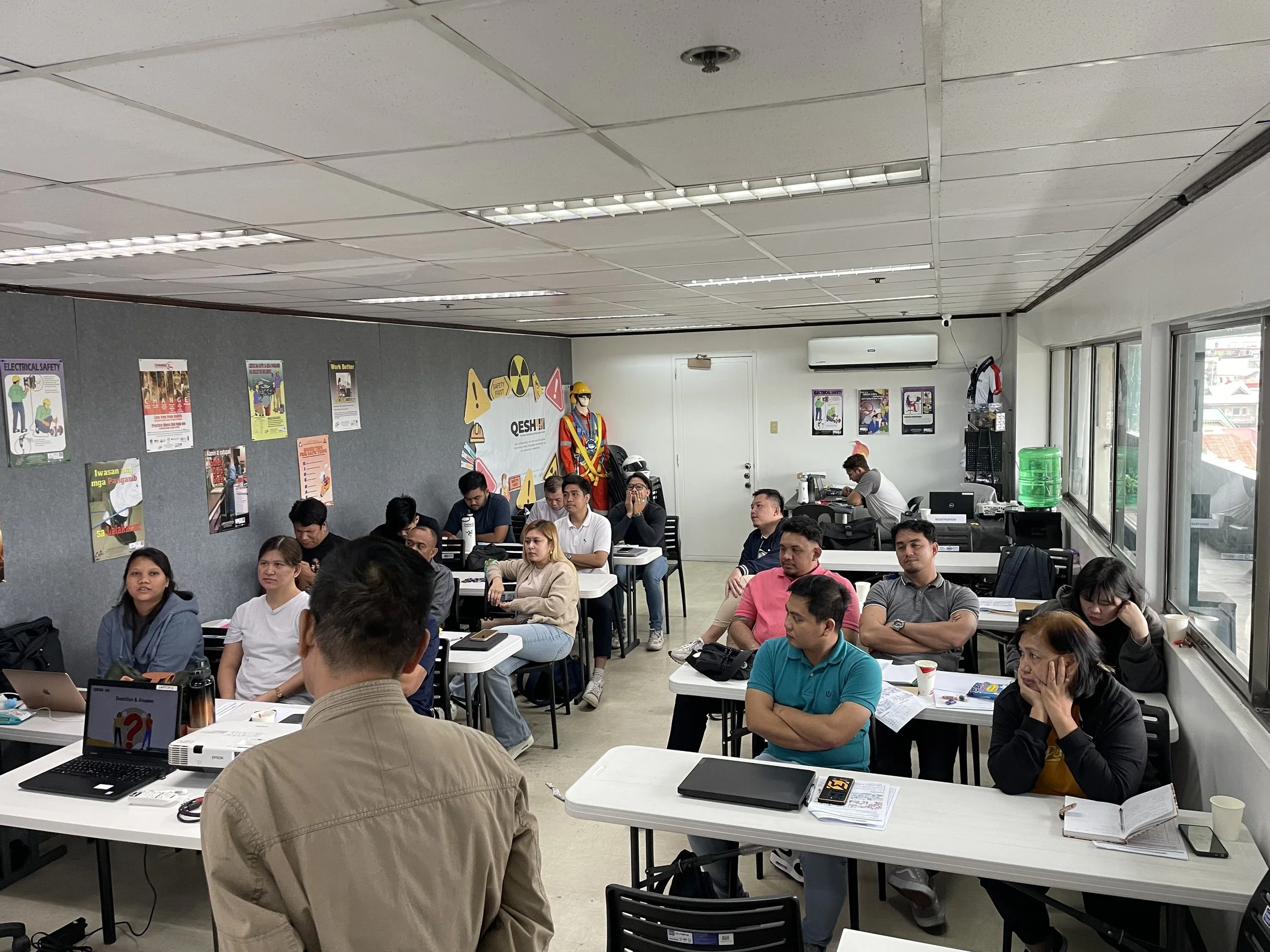 A classroom setting with adults seated at desks, listening to a presentation. The room has posters on the walls and a person is leading the session with a laptop and projector.