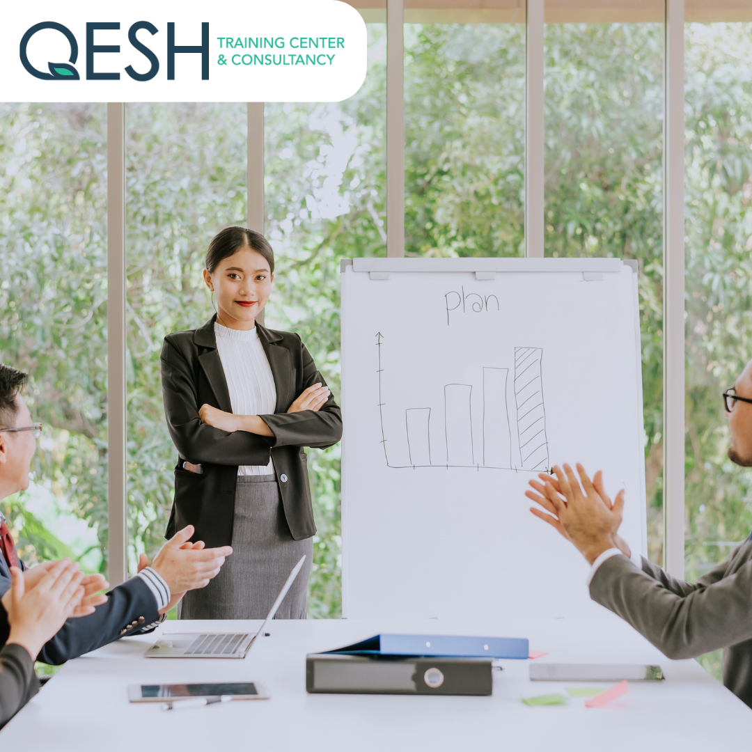 A businesswoman standing in front of a whiteboard with a bar graph, labeled 'plan,' while two colleagues applaud. The logo of QESH Training Center & Consultancy is visible. The setting is a modern office with large windows and greenery outside.